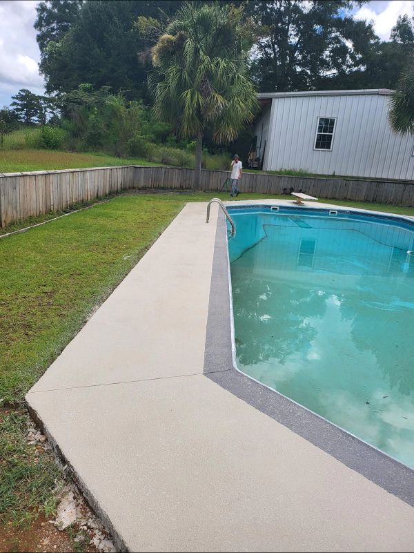 Poolside view of a concrete deck, a person in the distance, and a green lawn.