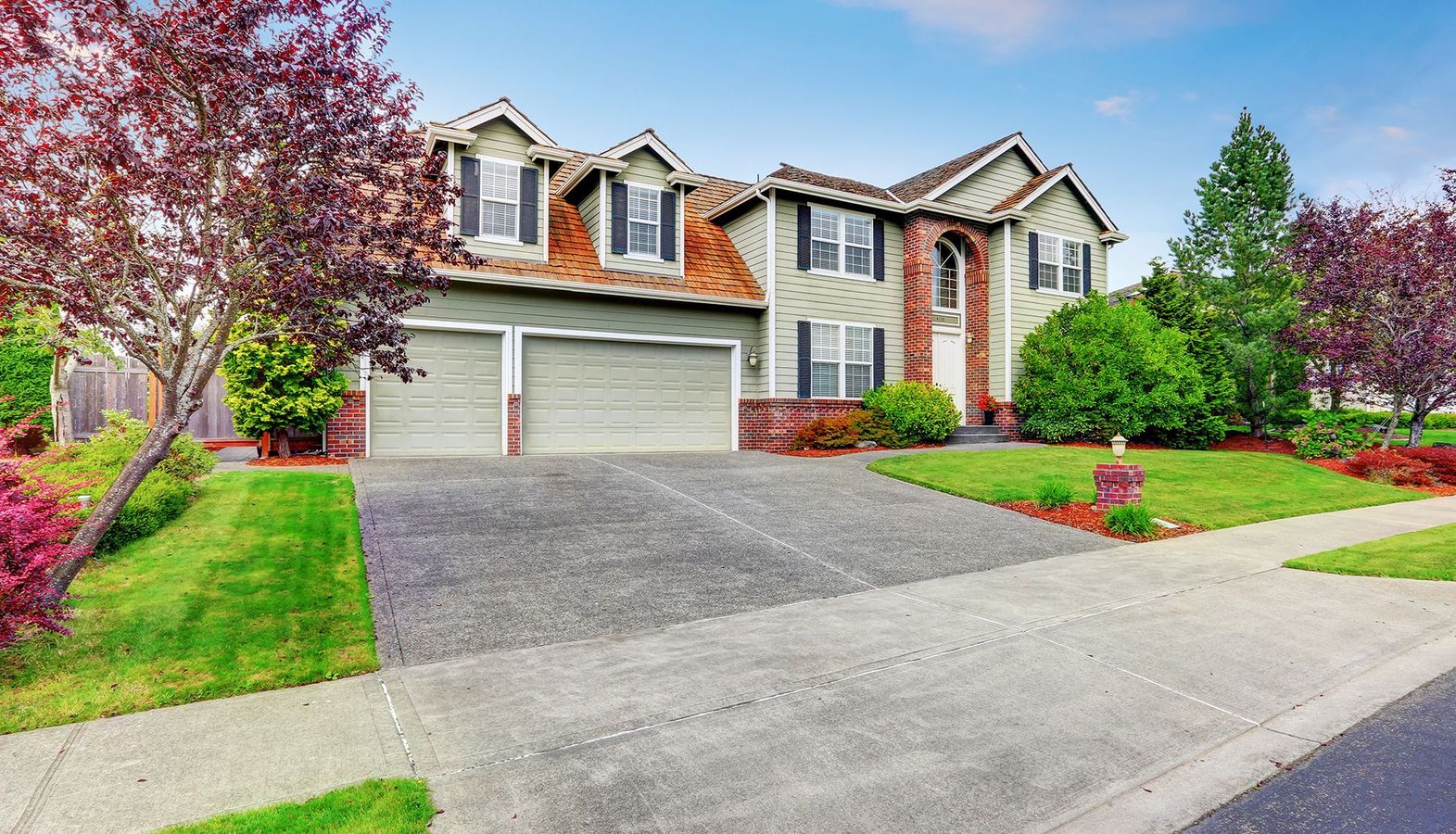 Large house with two-car garage, green siding, red brick accents, and a concrete driveway.