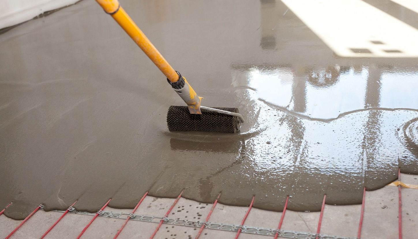 Worker smoothing wet cement on a floor with embedded red heating cables, using a brush tool.