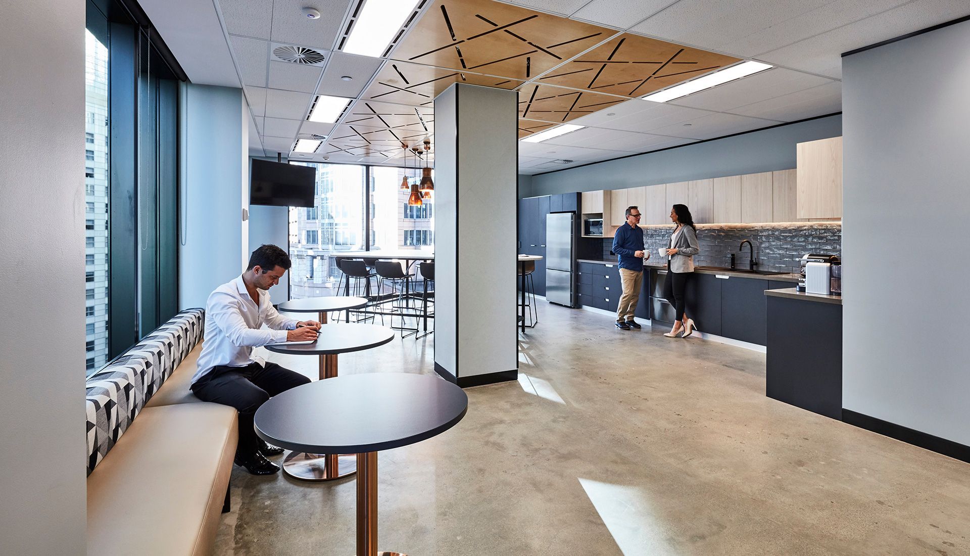 Office interior with a man at a table, a kitchen with people, and a long table by a window.