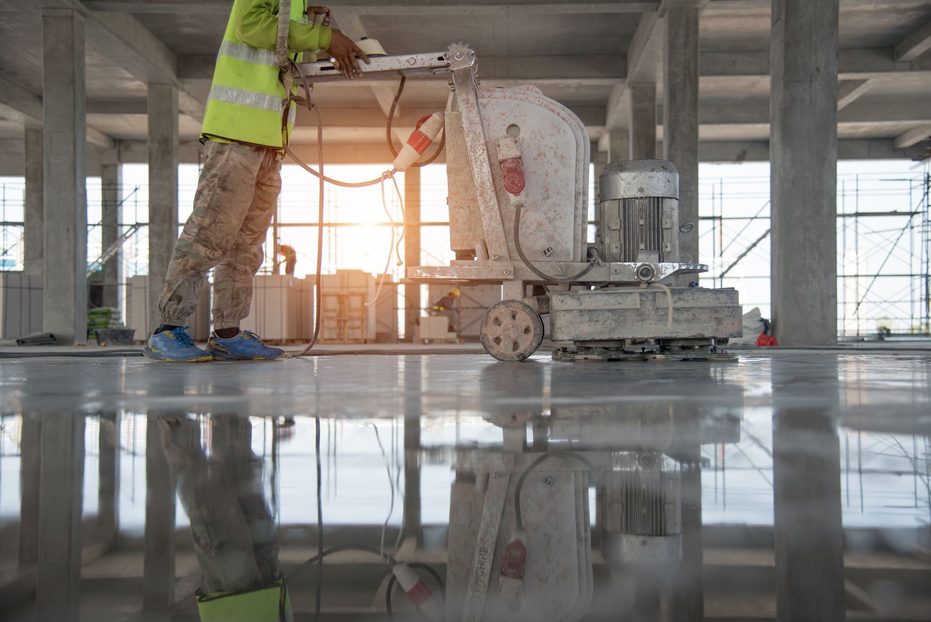 Worker using a floor grinder on a concrete surface inside a building under construction. Shiny floor reflects light.