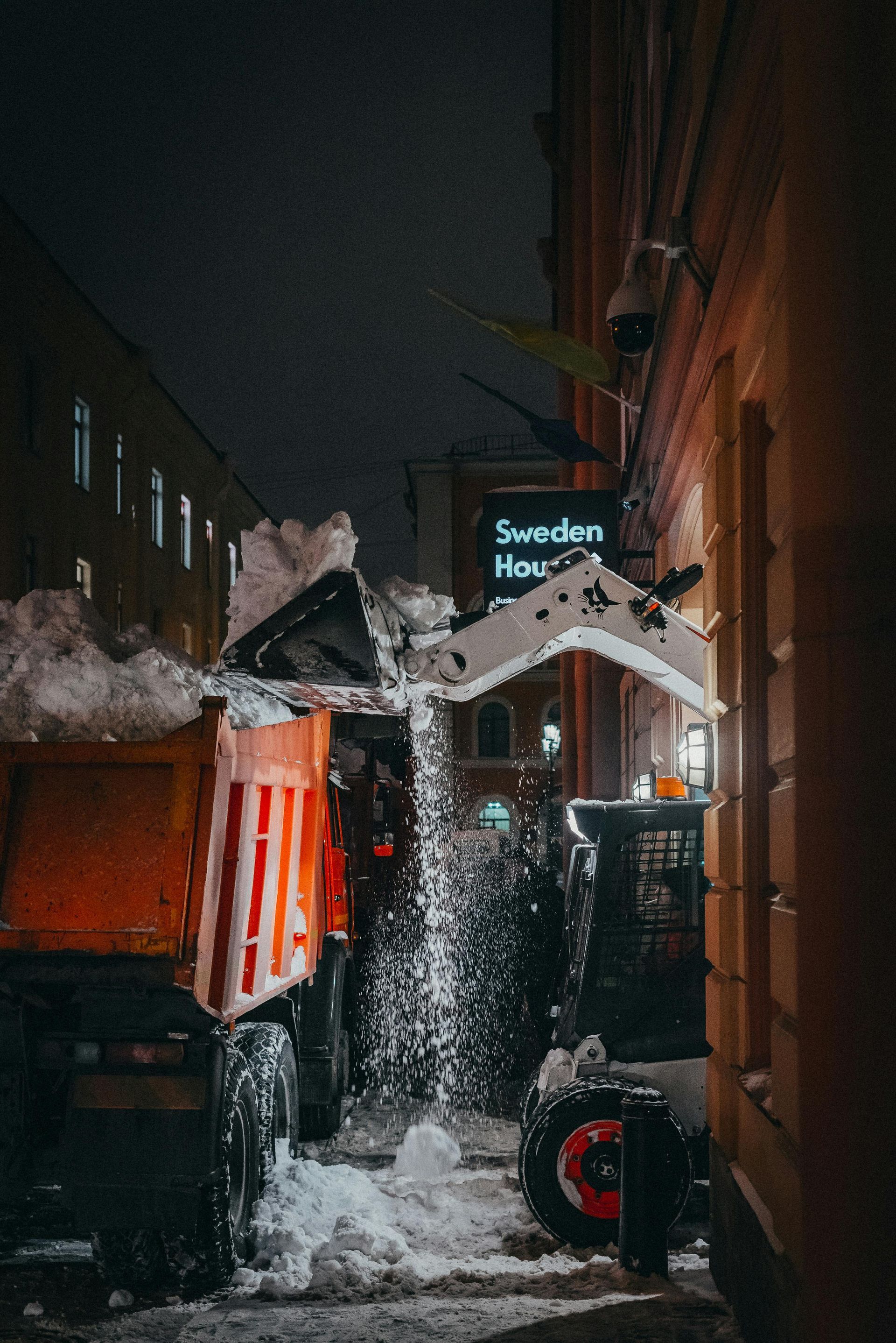 Snowplow dumping snow in a narrow street at night, next to a building with a sign.