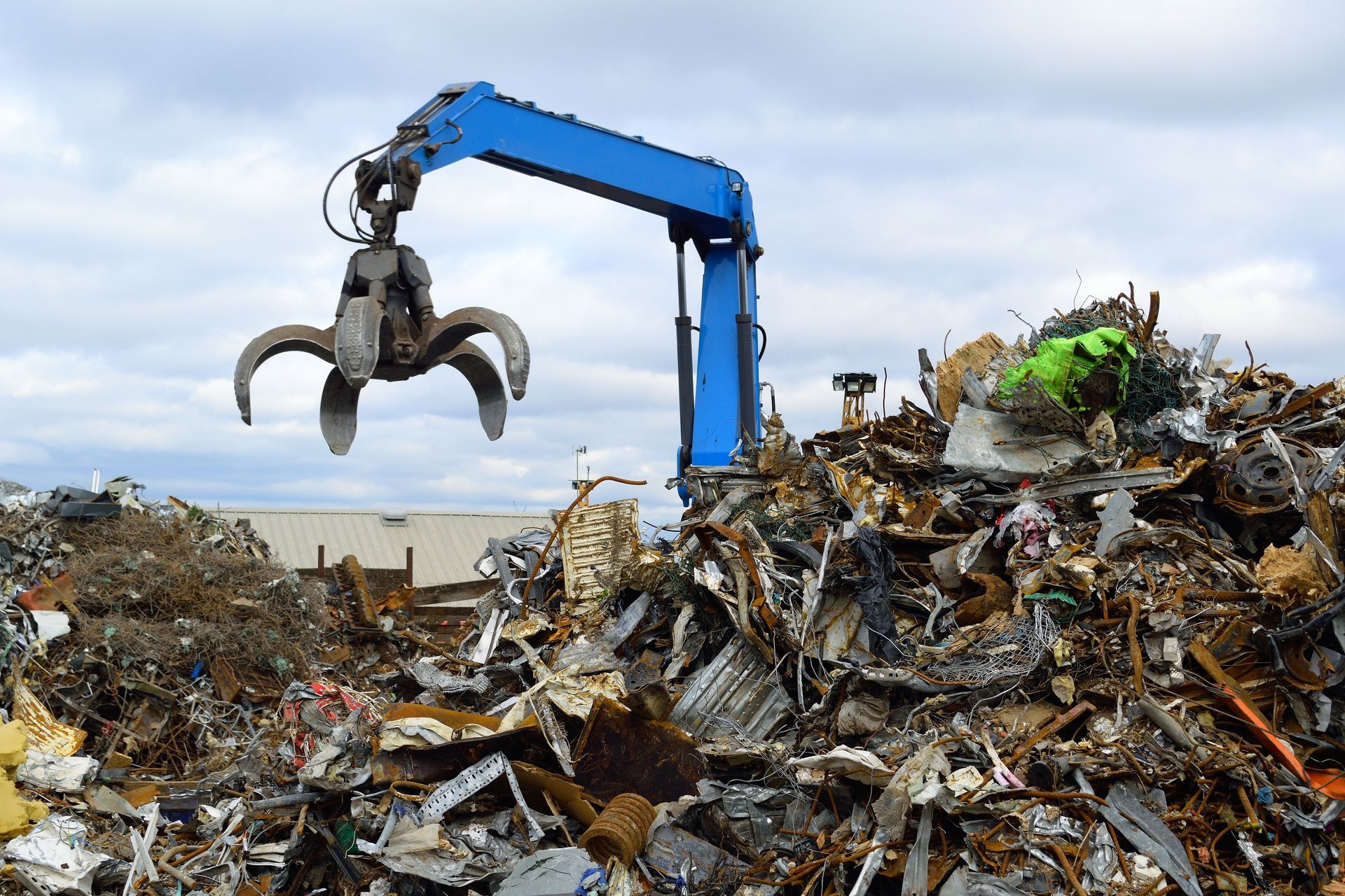 Blue claw crane lifting scrap metal in a junkyard. Cloudy sky in the background. Blue claw crane lifting scrap metal in a junkyard. Cloudy sky in the background.
