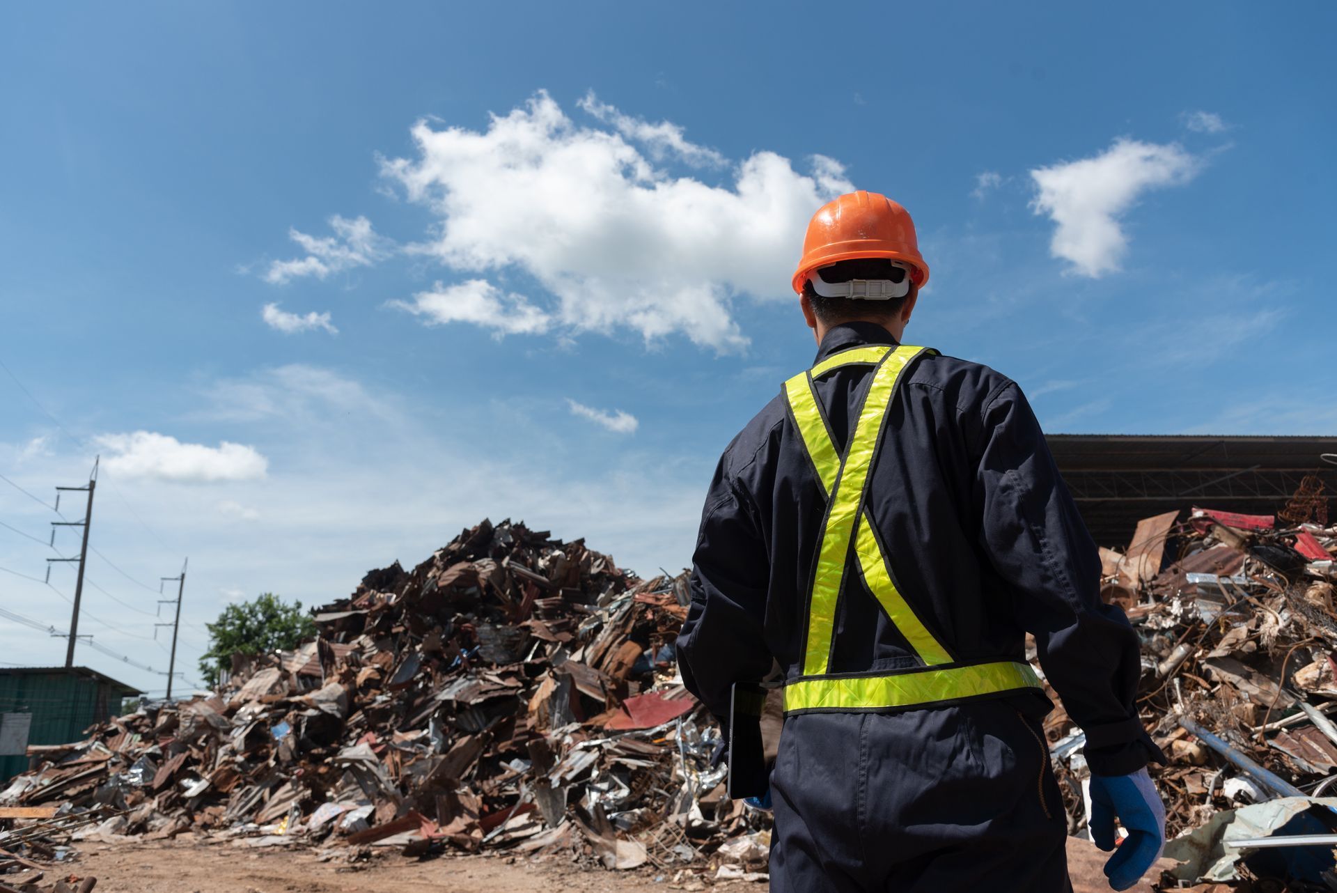 Worker in orange hard hat and safety vest, surveying a large pile of debris against a blue sky.