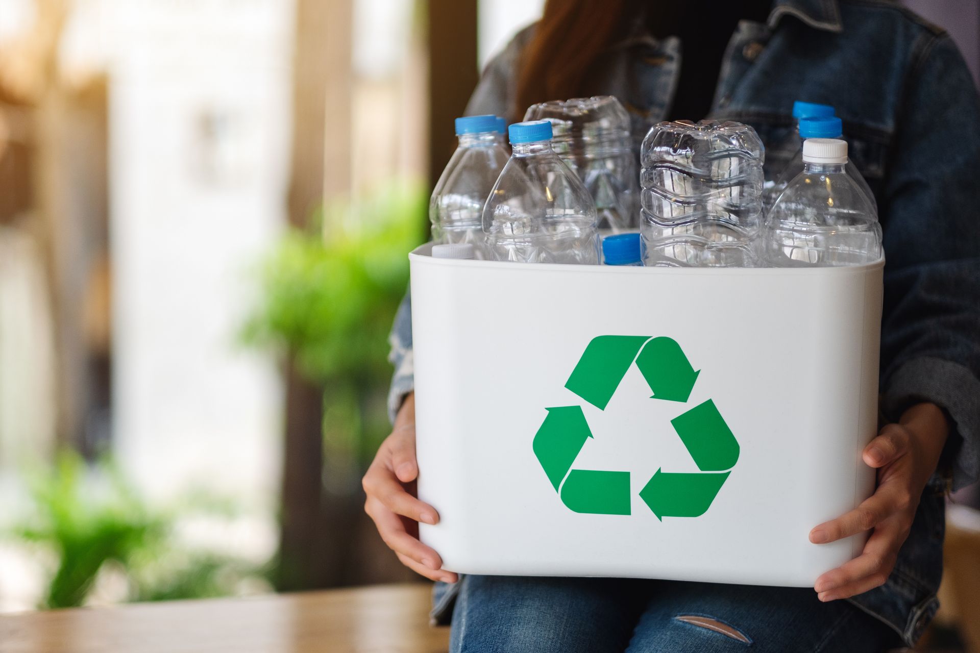 Person holding a white recycling bin filled with clear plastic water bottles, green recycling symbol on the bin.