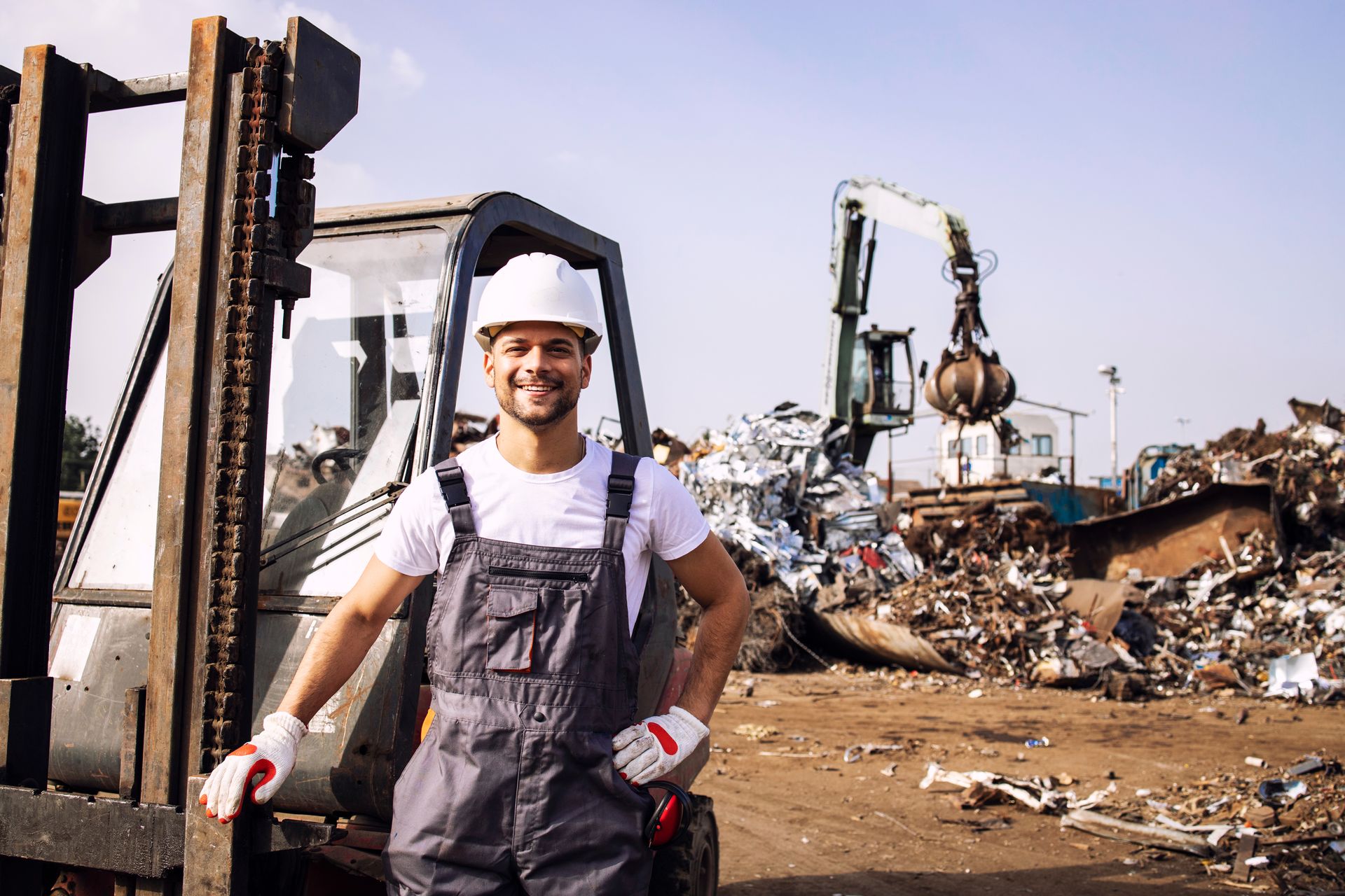 Smiling worker in hard hat and overalls by forklift, scrap yard with machinery in background.