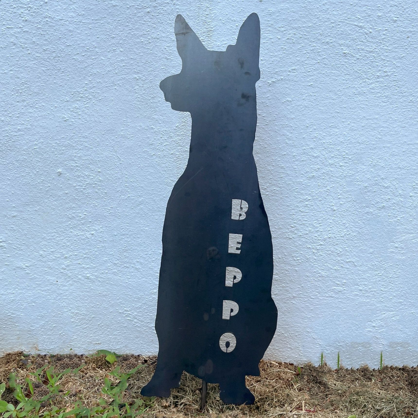 Schwarze Metall-Hundesilhouette mit dem Namen „Beppo“ in silbernen Buchstaben. Sitzt vor einer weißen Wand.
