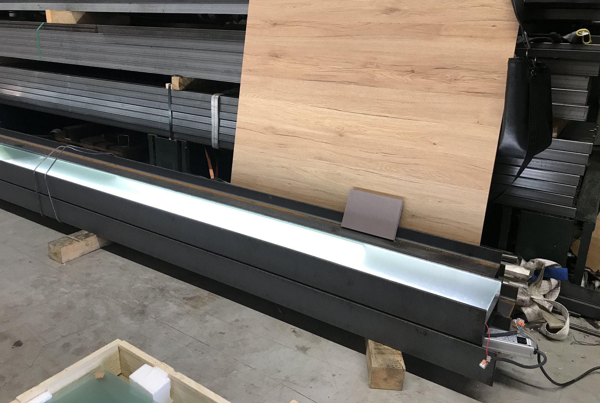 Lange, dunkle Metallkonstruktion mit beleuchtetem Innenraum, möglicherweise Beleuchtungseinrichtungen, vor einem Holzhintergrund.