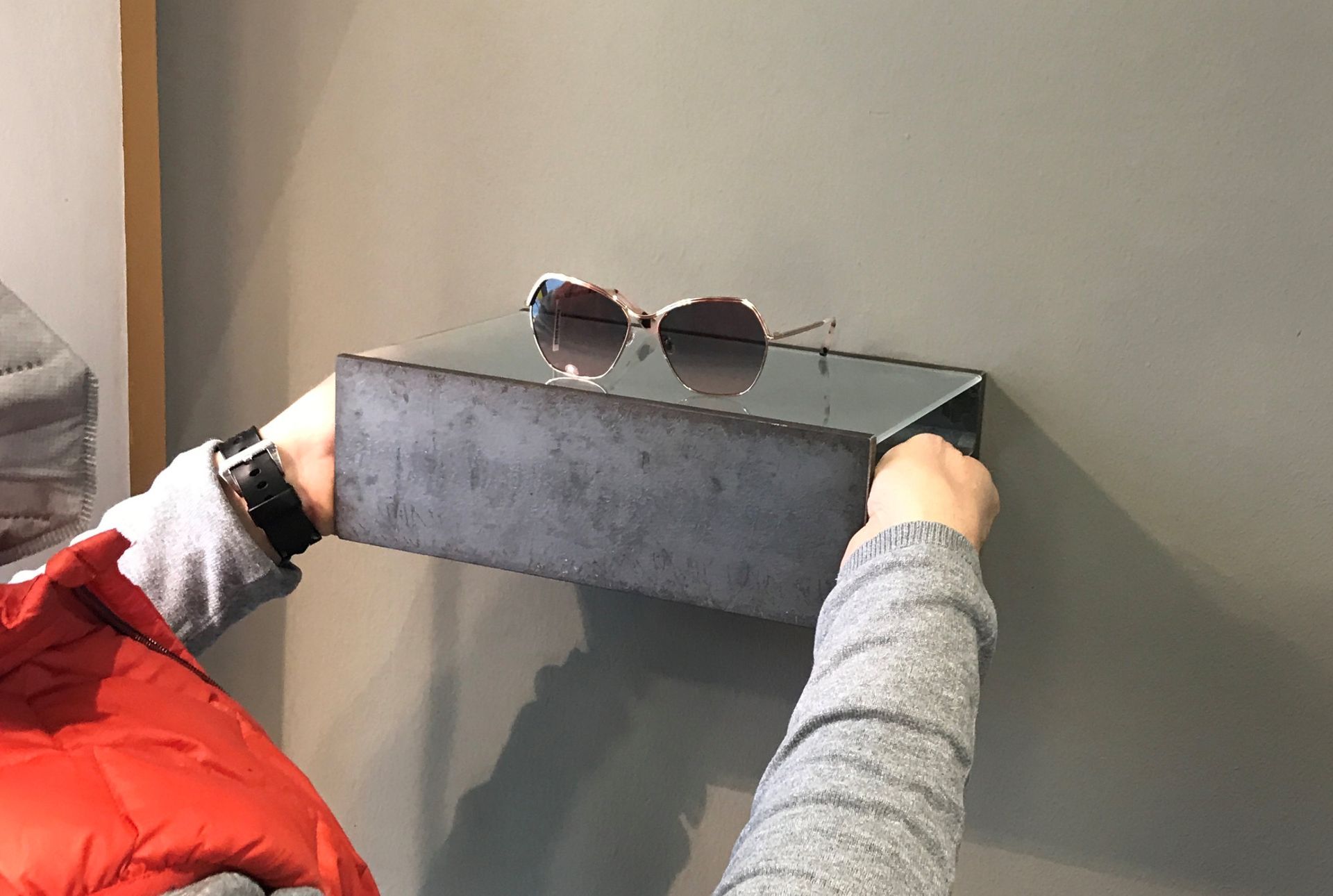 Eine Person hält ein graues Regal mit Sonnenbrillen an einer Wand.