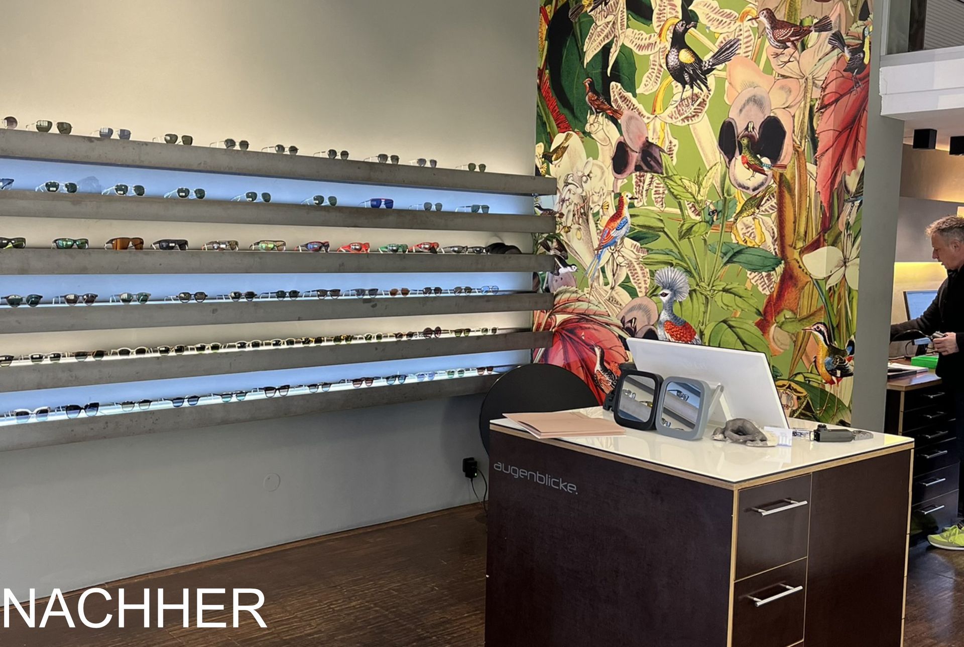 Innenansicht eines Optikergeschäfts mit Brillen in Ausstellungsregalen, einem Empfangstresen und einem floralen Wandbild.