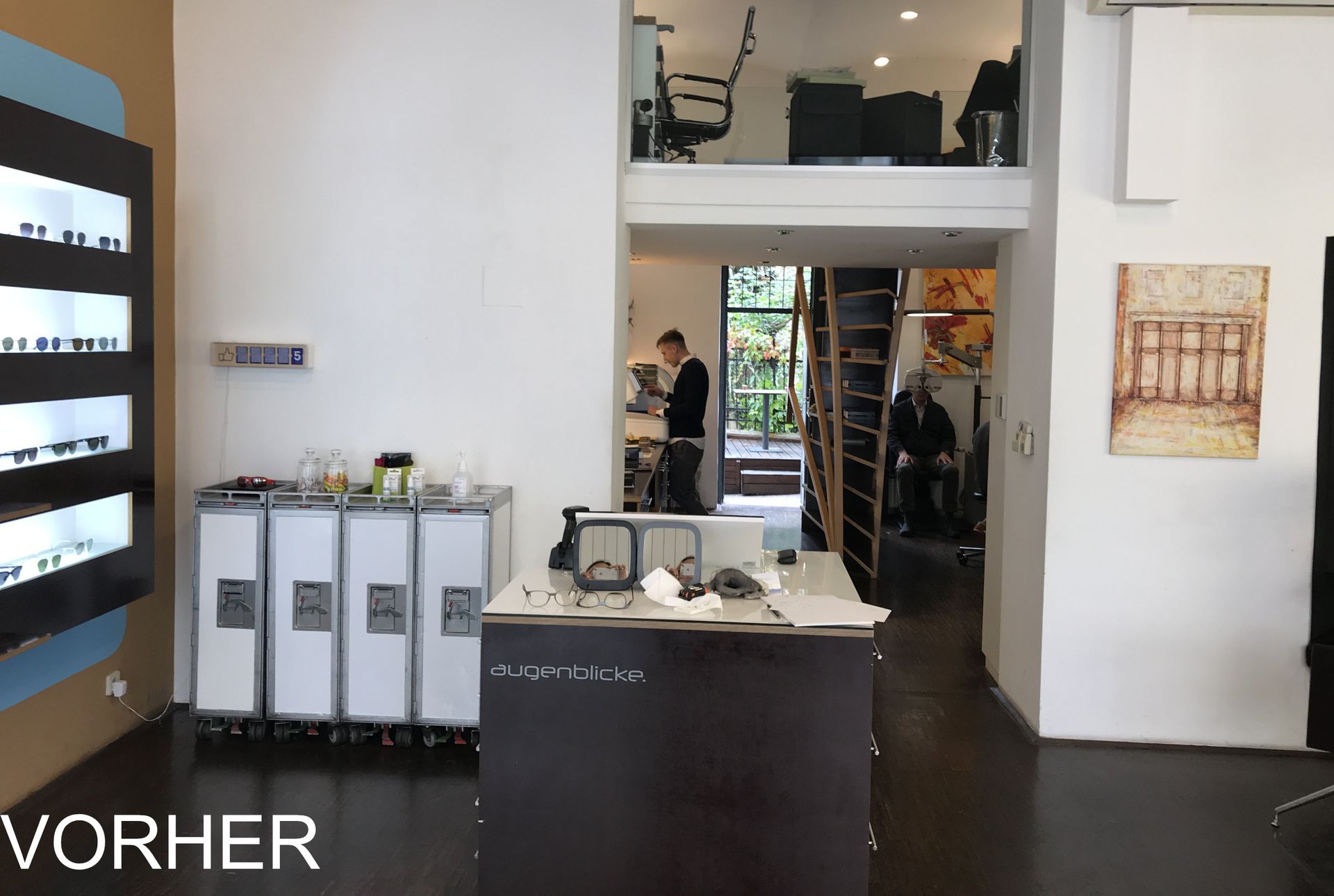 Innenbereich eines Ladengeschäfts mit Brillenauslagen. Eine Person steht im Türrahmen eines zweiten Stockwerks.