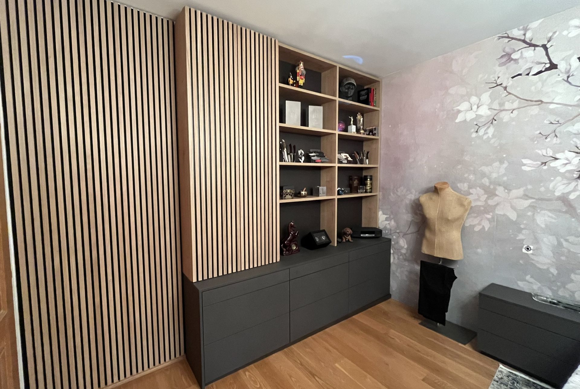 Aufbewahrungsmöbel aus Holz mit vertikalen Lamellen, offene Regale mit Dekorationsartikeln, graue Schränke, florale Tapete, Schaufensterpuppe.