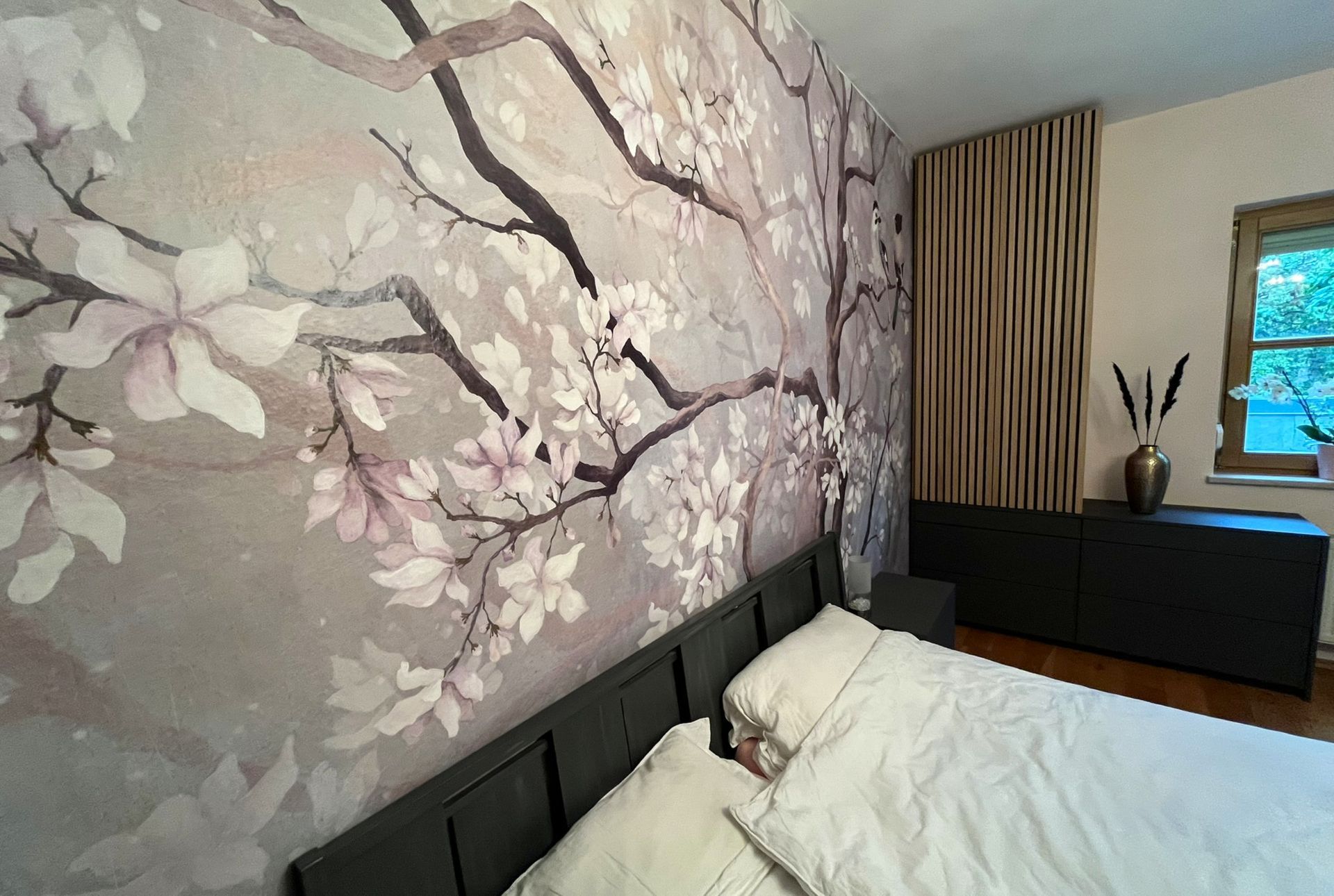 Schlafzimmer mit floralem Wandbild, Bett, schwarzem Sideboard und dekorativer Holzvertäfelung.