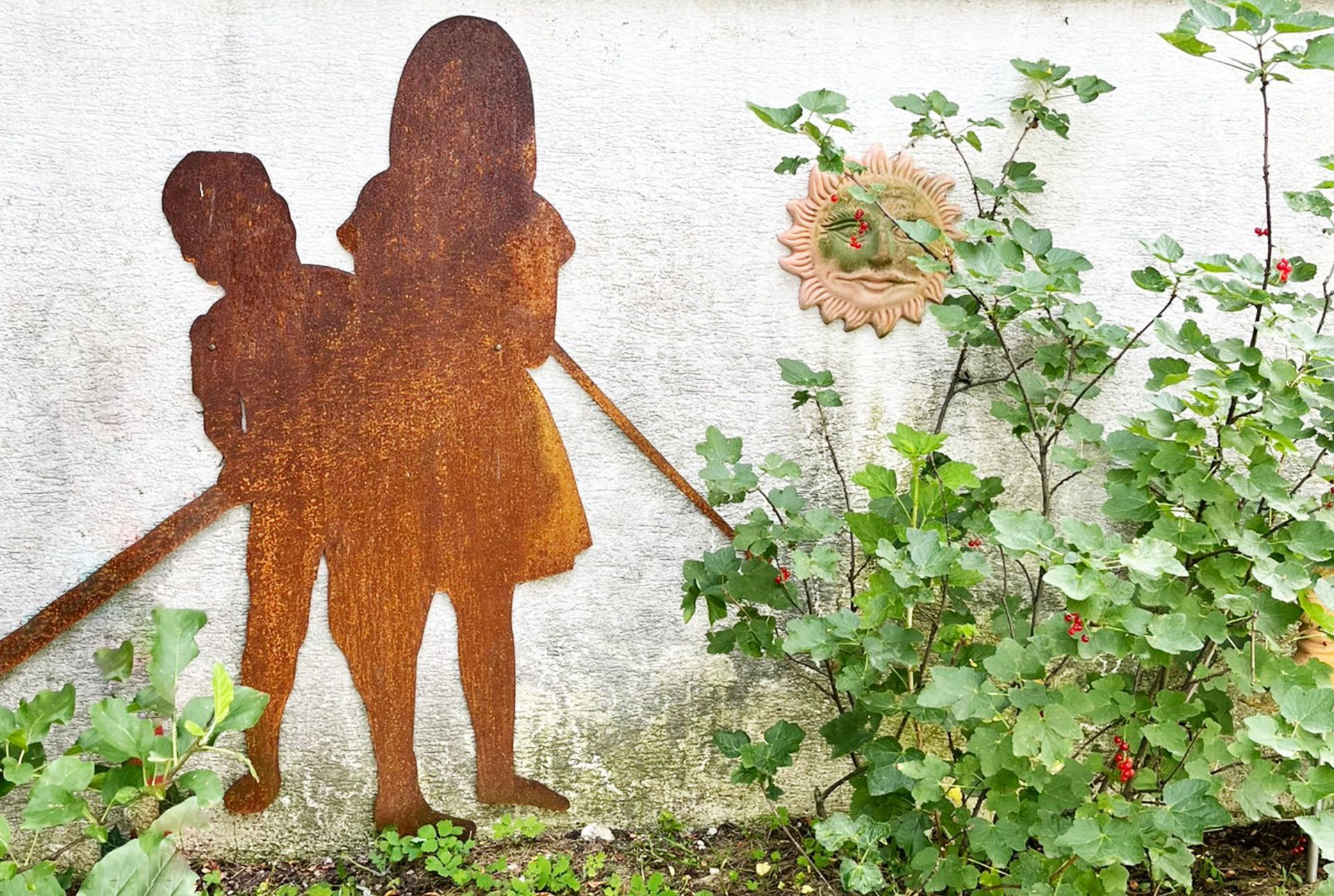 Rostige Metallsilhouette zweier Figuren neben Pflanzen an einer weißen Wand.