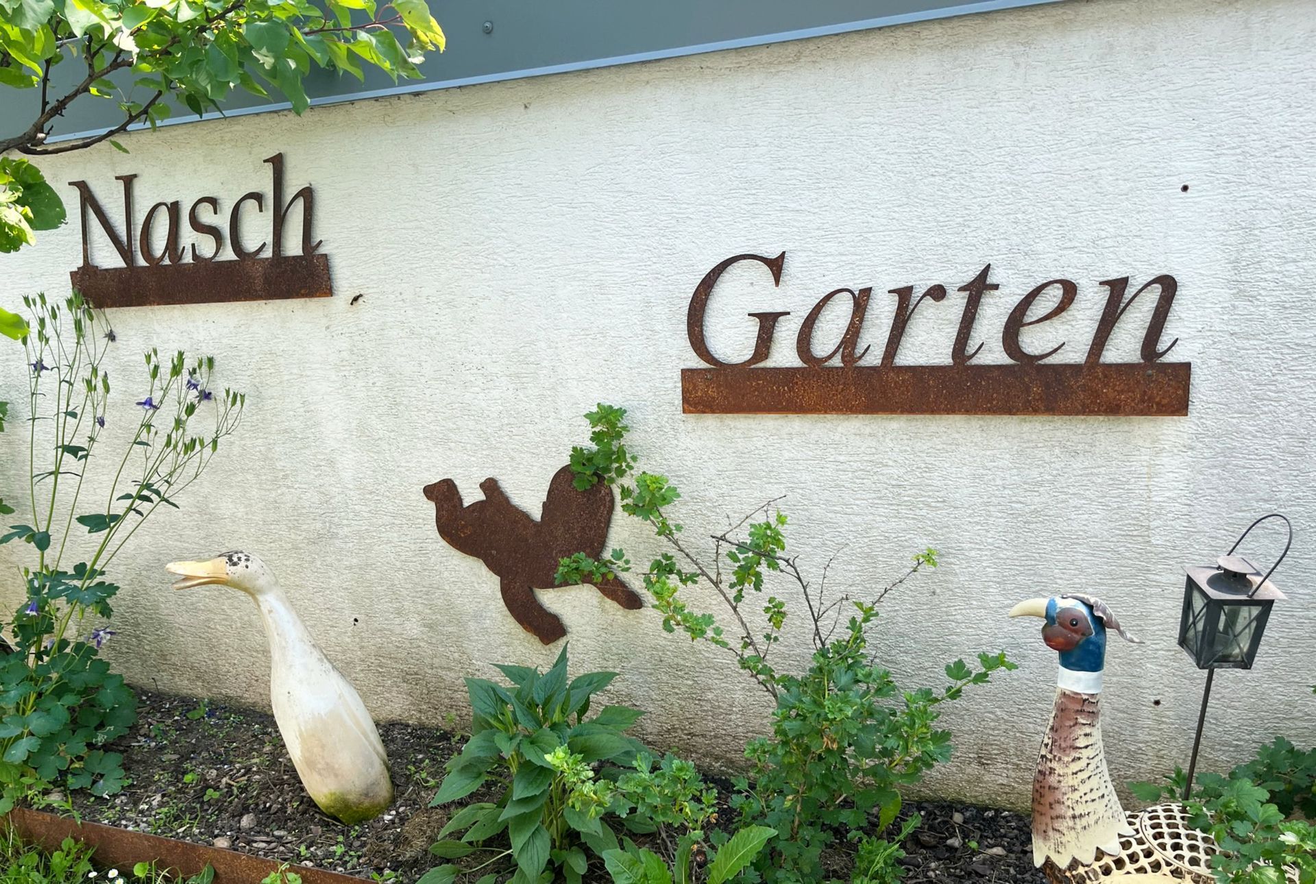 Ein Schild mit der Aufschrift „Nasch Garten“ an einer Stuckwand mit Metallverzierungen von Vögeln und Grünpflanzen.