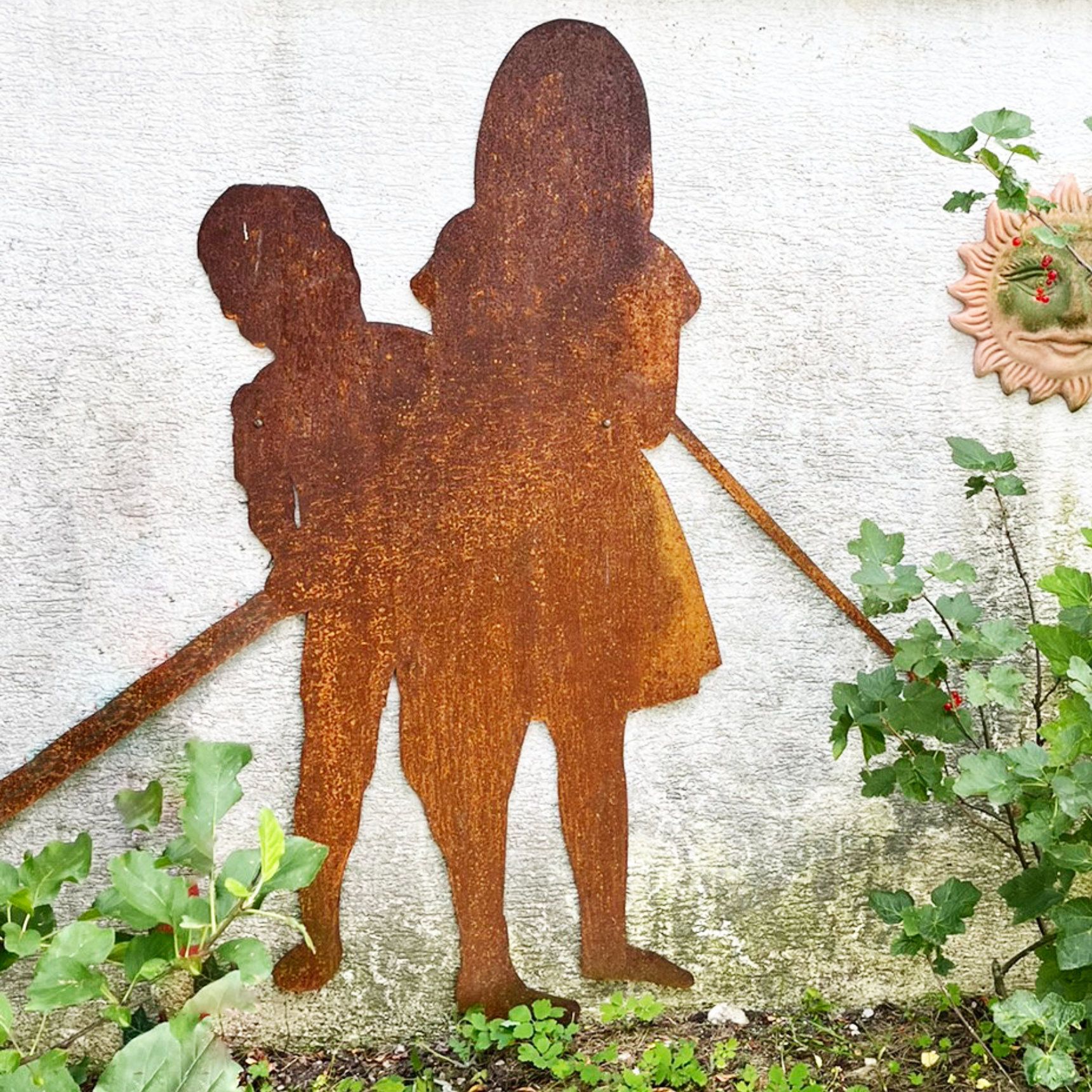 Die rostige Metallsilhouette zweier Kinder an einer Wand, die einen Stock halten, in der Nähe Grünpflanzen.