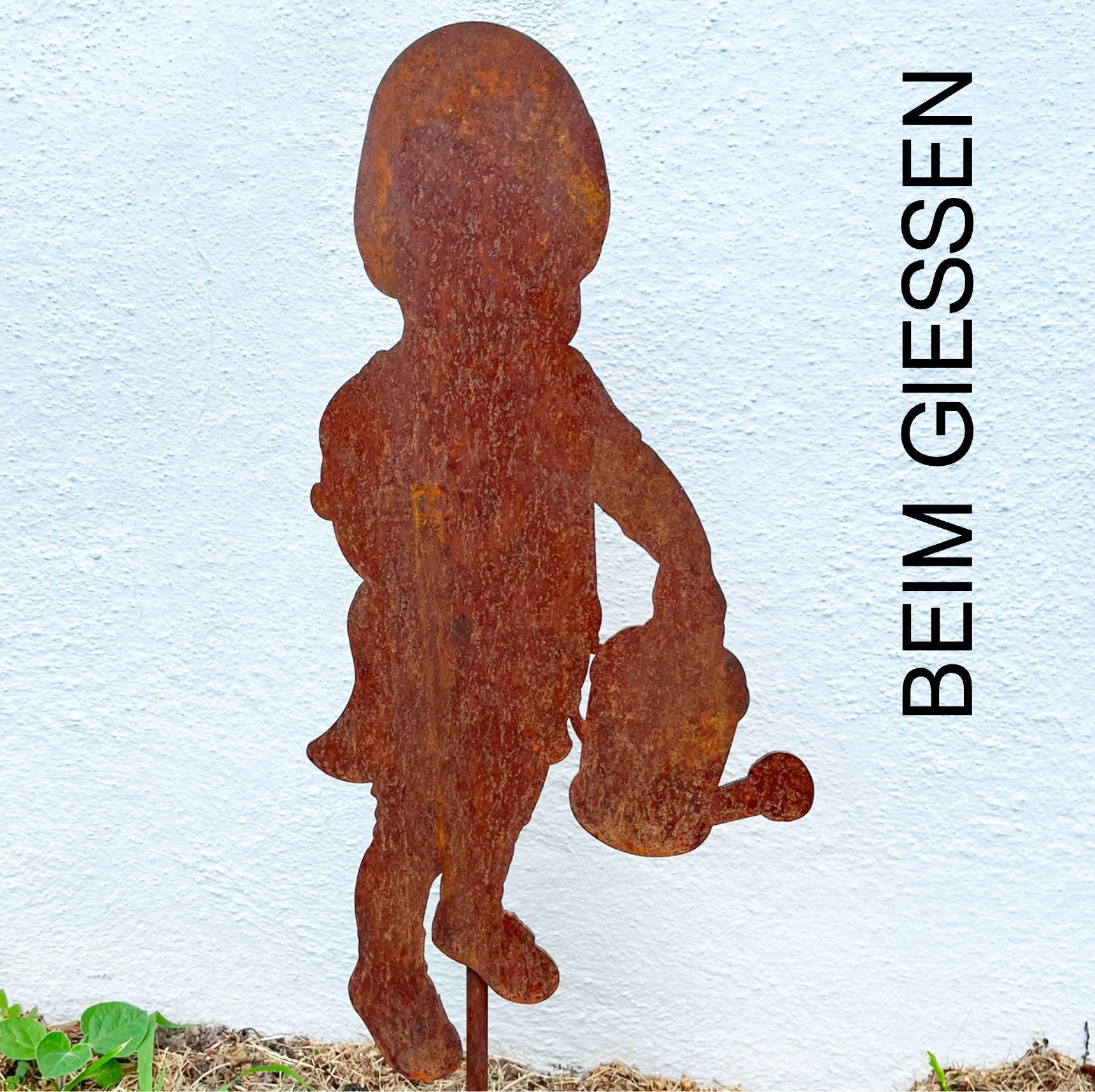 Rostige Metallsilhouette eines Kindes mit einer Gießkanne in einem Garten; Text „BEIM GIESSEN“.