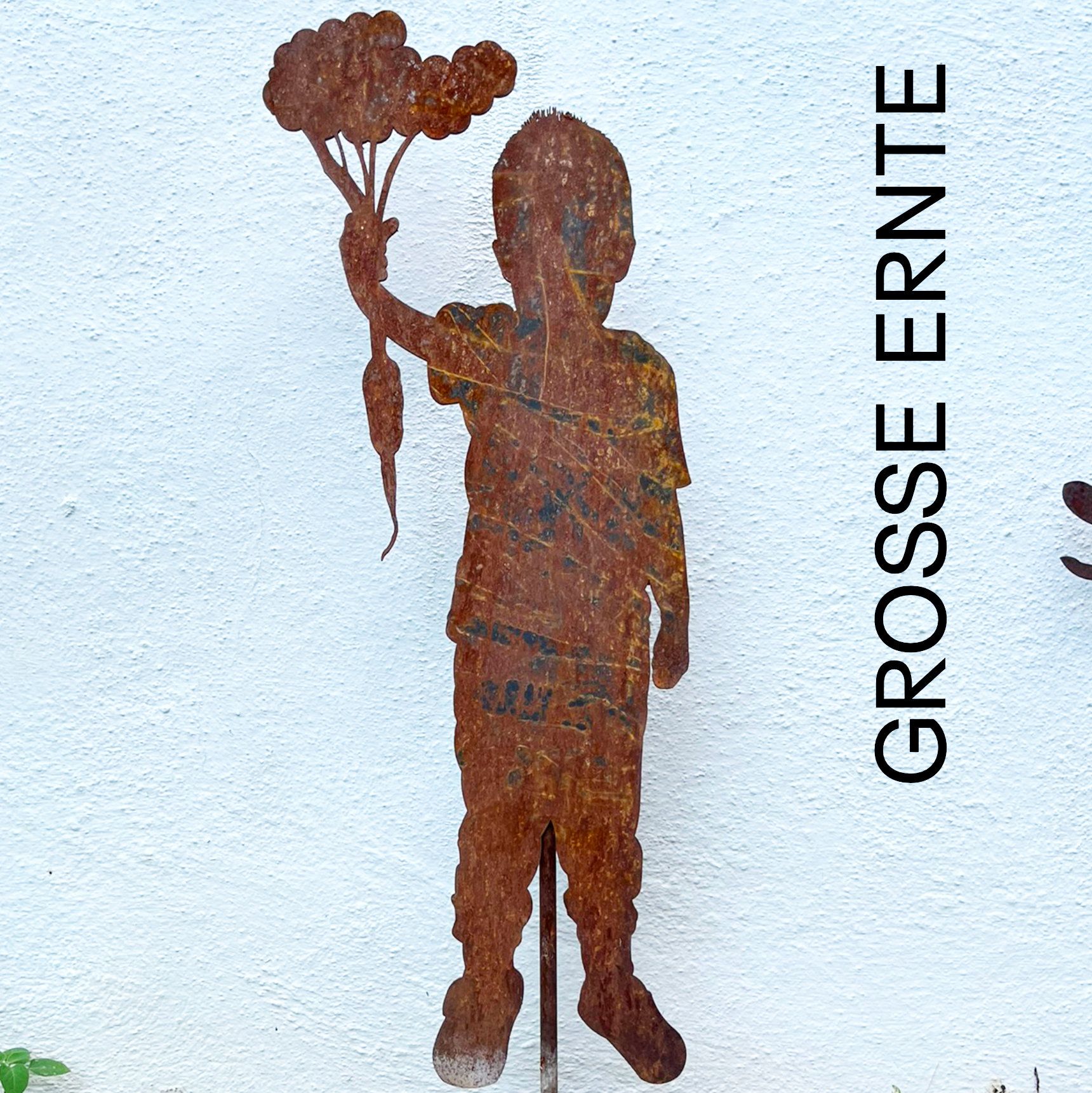 Rostige Metallsilhouette eines Kindes mit einer Karotte. Weißer Wandhintergrund mit dem Schriftzug „GROSSE ERNTE“.