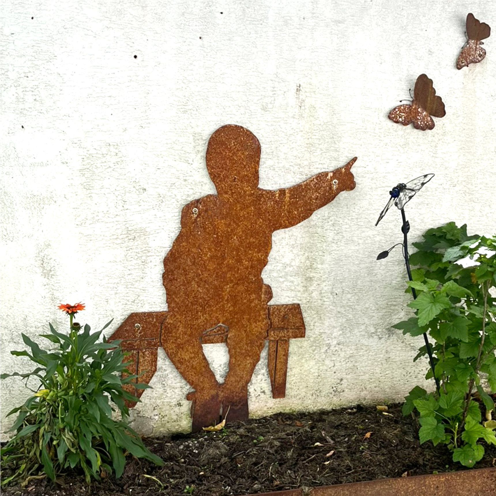Die Silhouette einer Person aus rostigem Metall, die auf einer Bank sitzt und zeigt, daneben Schmetterlinge 
