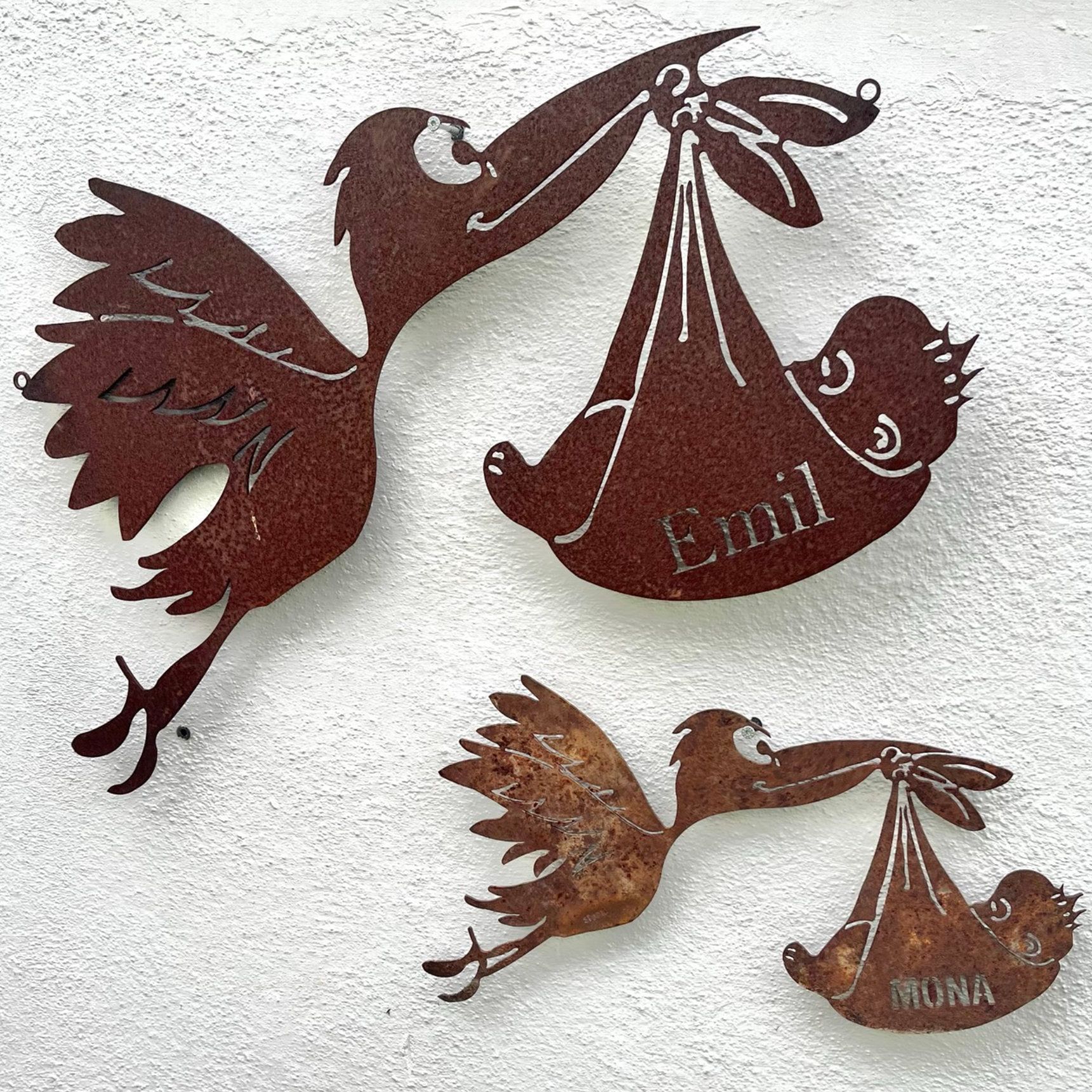 Metallene Storchenfiguren, die Babys tragen, zwei davon mit Namen: Emil und Luna, an einer weißen Wand.