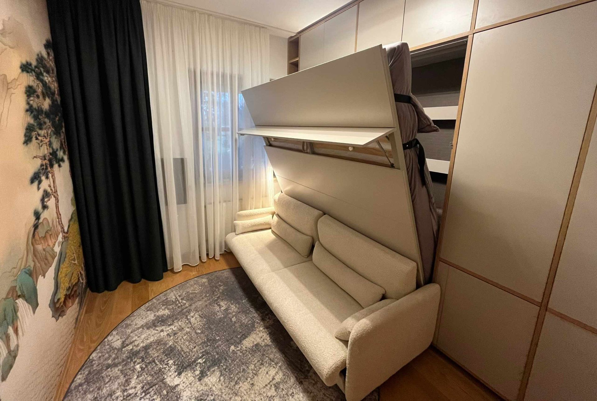 Ein Sofa, das sich in einem kompakten Zimmer neben einem Fenster und einem Kleiderschrank in ein Wandbett verwandeln lässt.