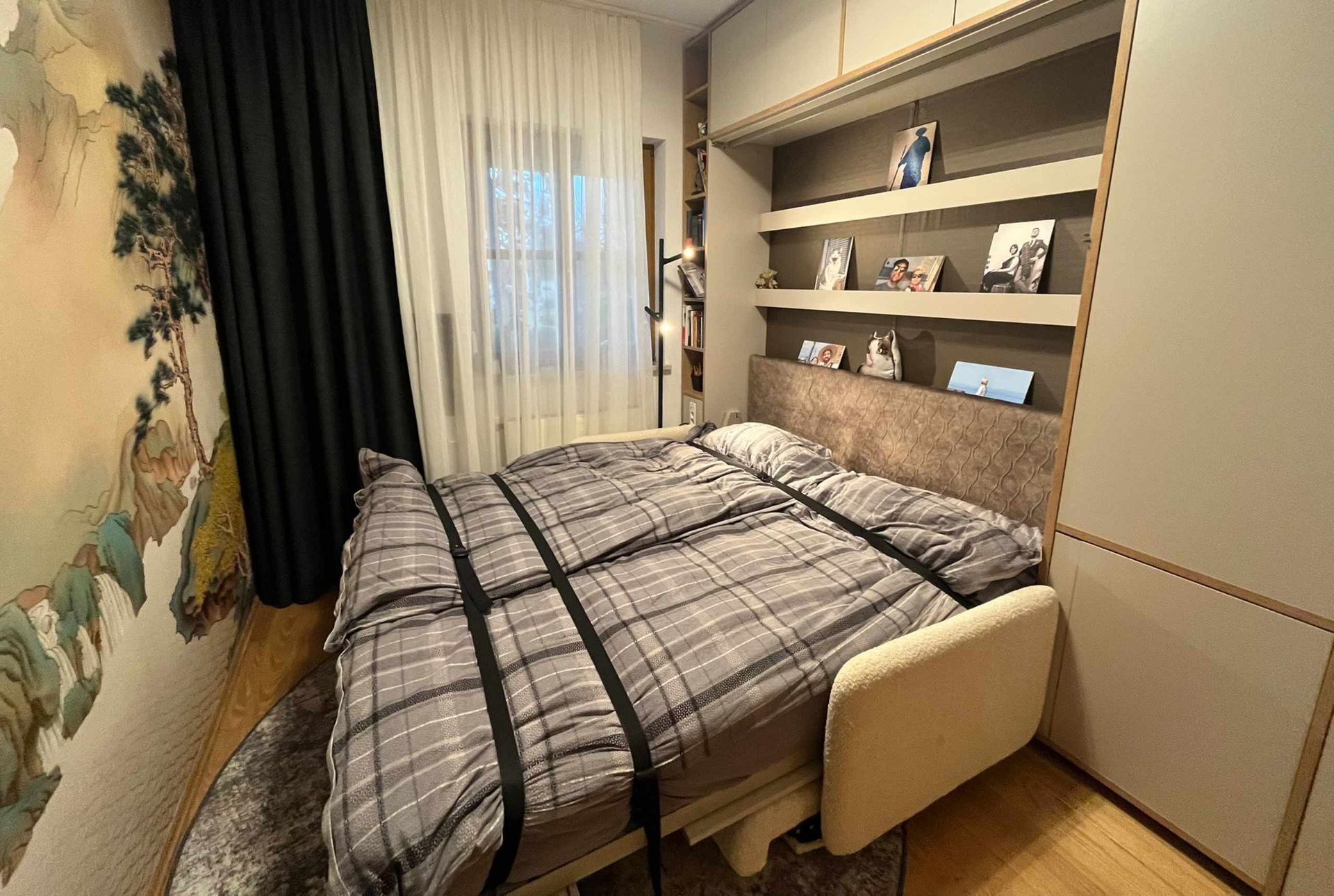 Schlafzimmer mit Klappbett. Graue, karierte Bettwäsche, mit Gurten befestigt; Einbauregale und Kleiderschrank. Asiatisches Wandbild.