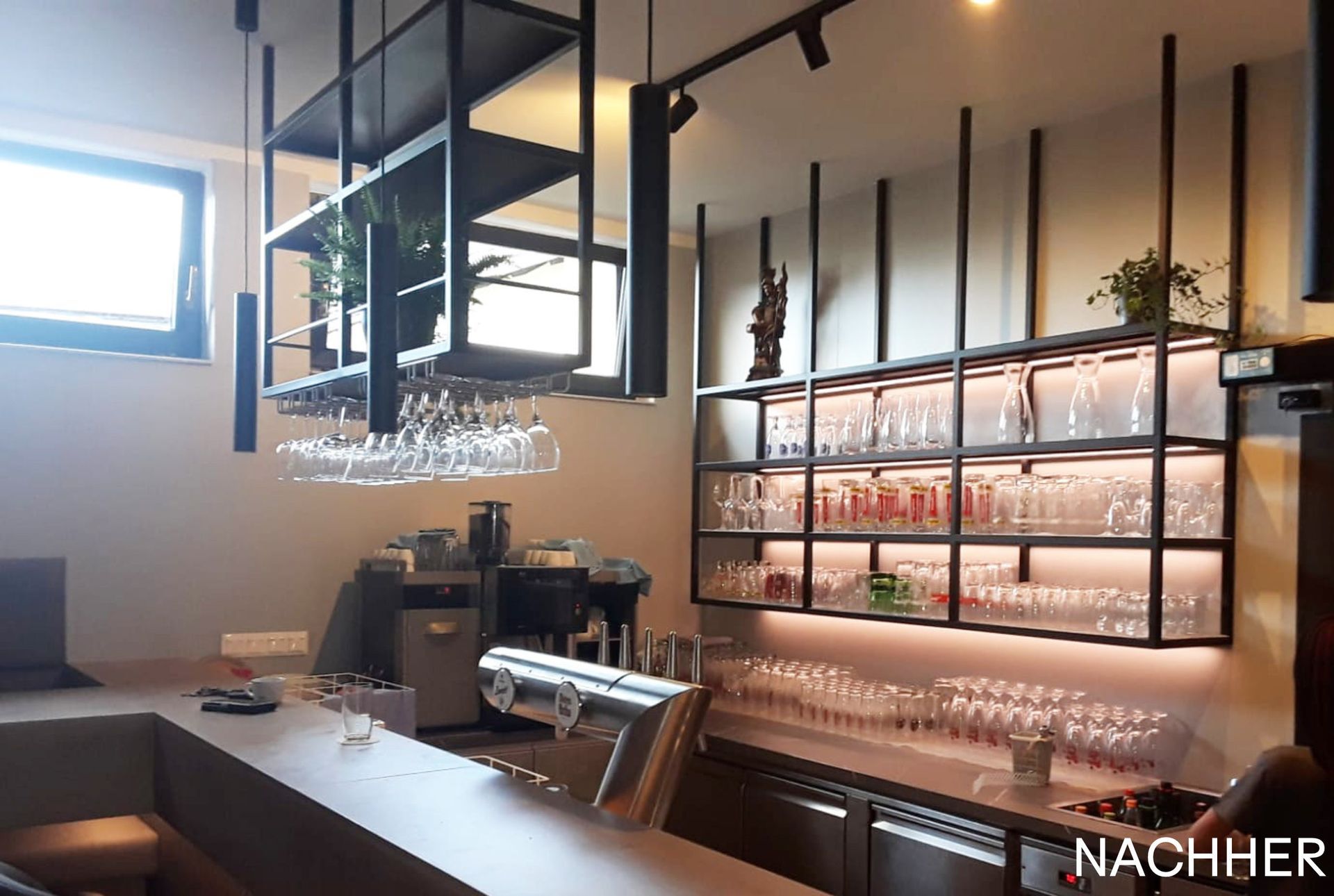 Bar-Interieur mit Metallregalen, Gläsern und Pflanzen, beleuchtet mit warmem Licht.