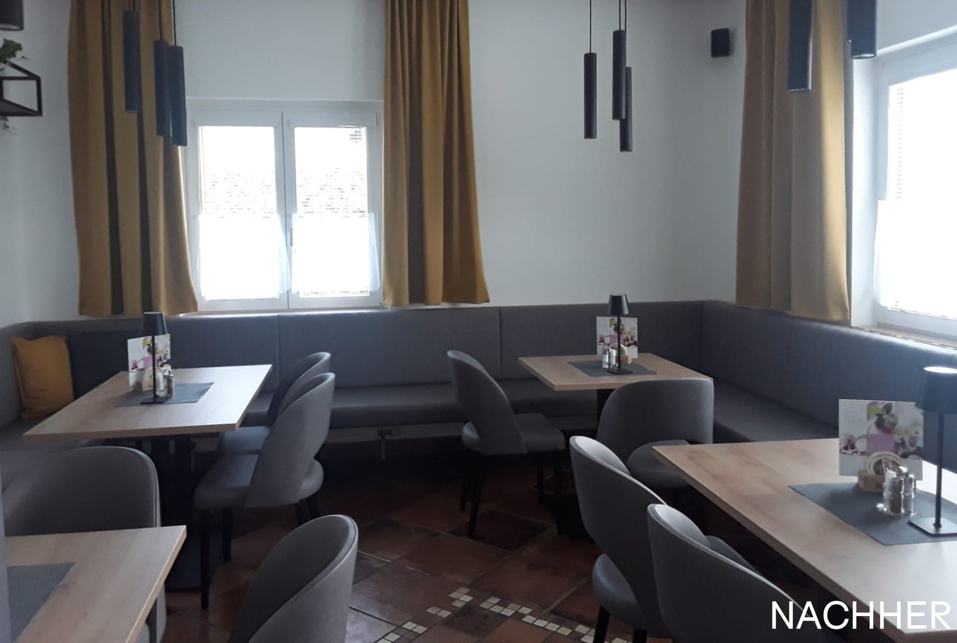 Restaurantinterieur: graue Sitznischen, Tische, Stühle, Fenster mit goldenen Vorhängen und dunkle Pendelleuchten.