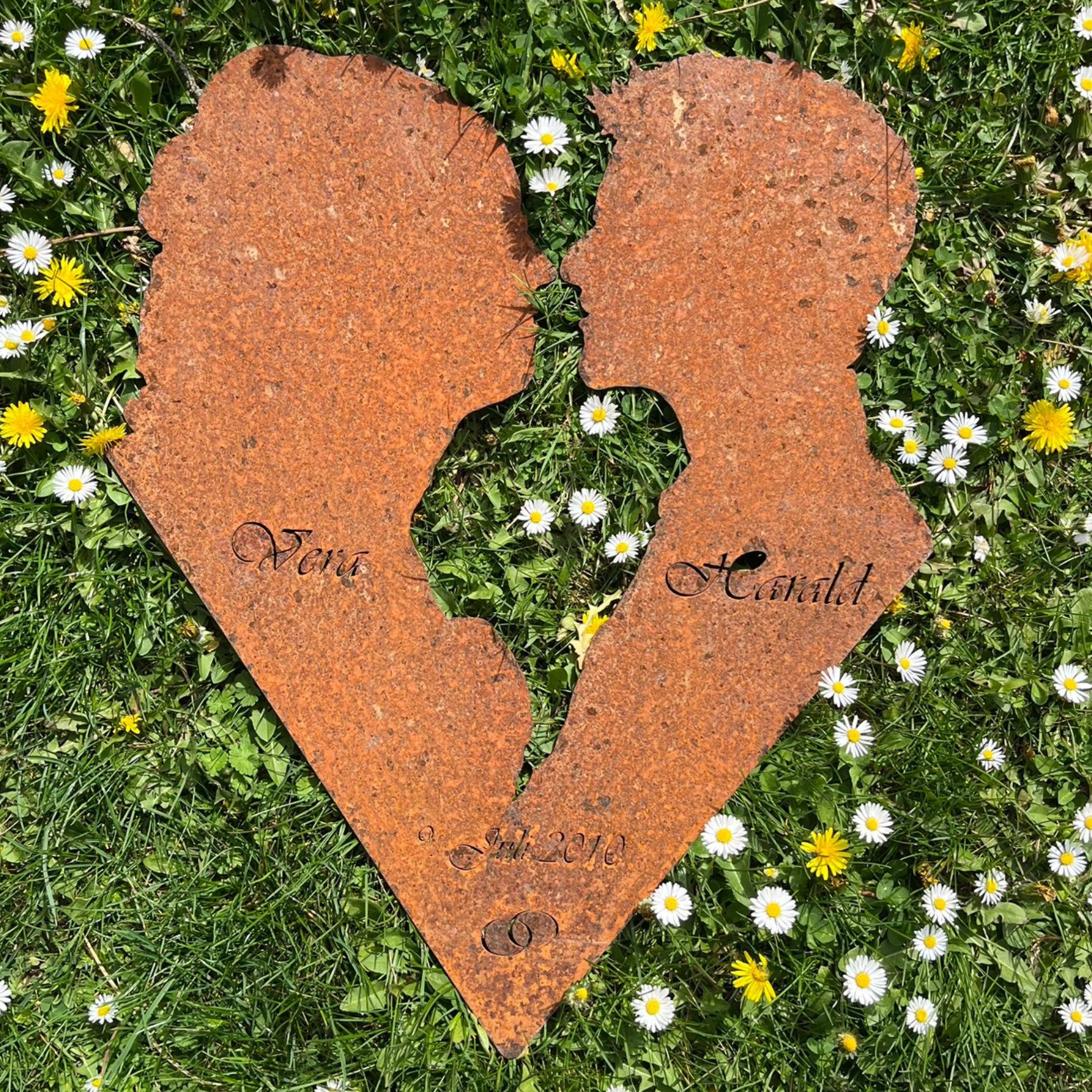 Zwei rostige Metallsilhouetten von Menschen in Form eines gebrochenen Herzens, mit Namen und Datum auf Gras.