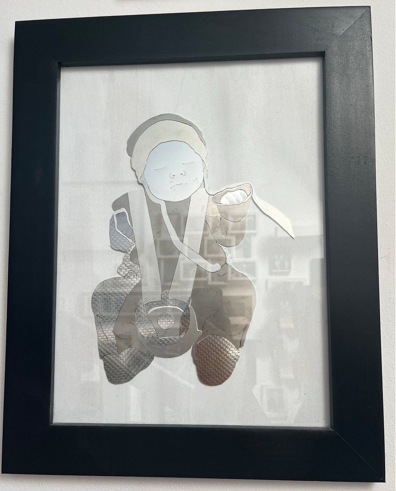 Gerahmtes Kunstwerk einer sitzenden Figur aus Spiegelglas auf weißem Hintergrund mit dunkelschwarzem Rahmen.