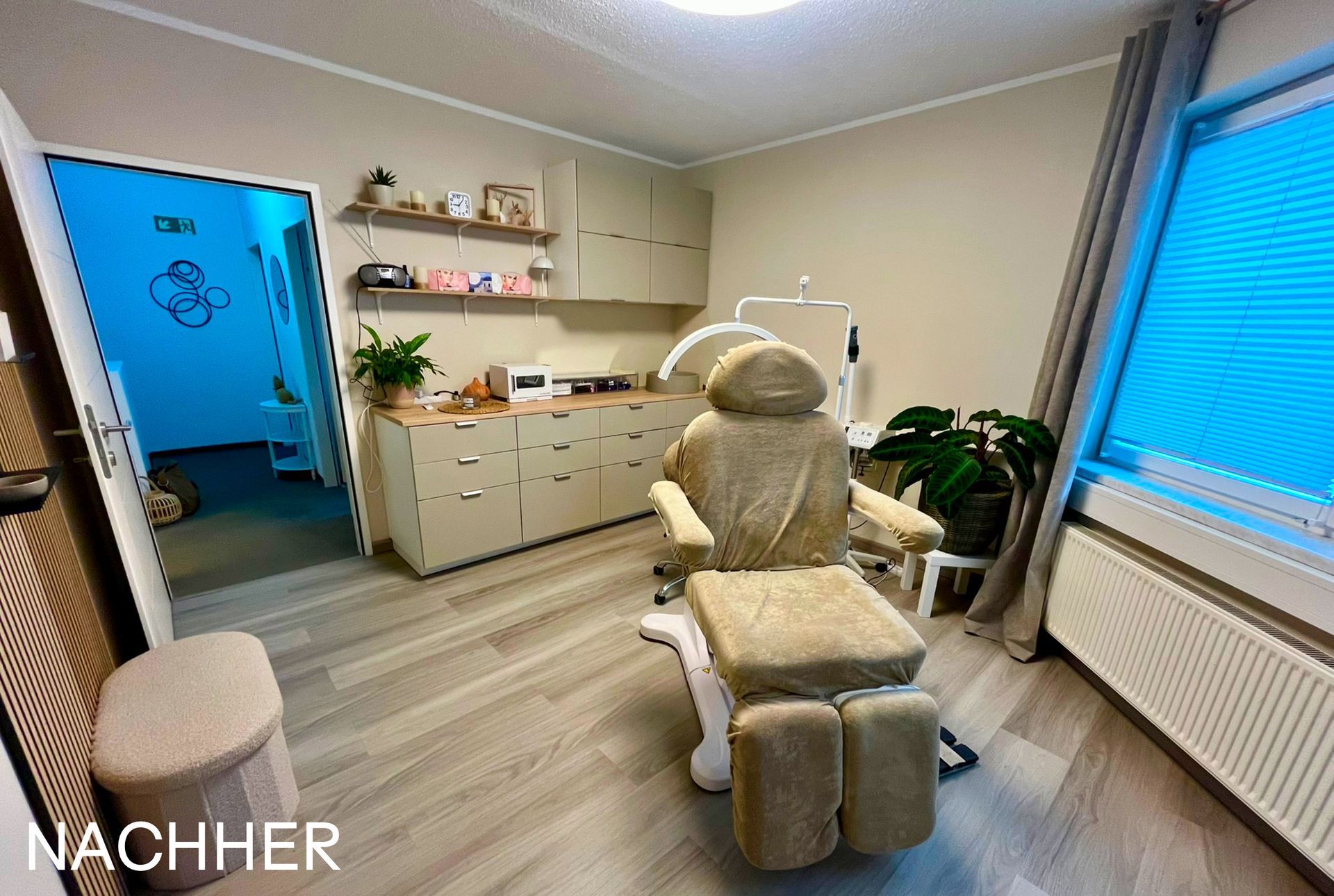 Innenansicht einer beigefarbenen Arztpraxis oder eines Spas mit Behandlungsstuhl, Schränken und einer Tür, die zu einem blau beleuchteten Bereich führt.