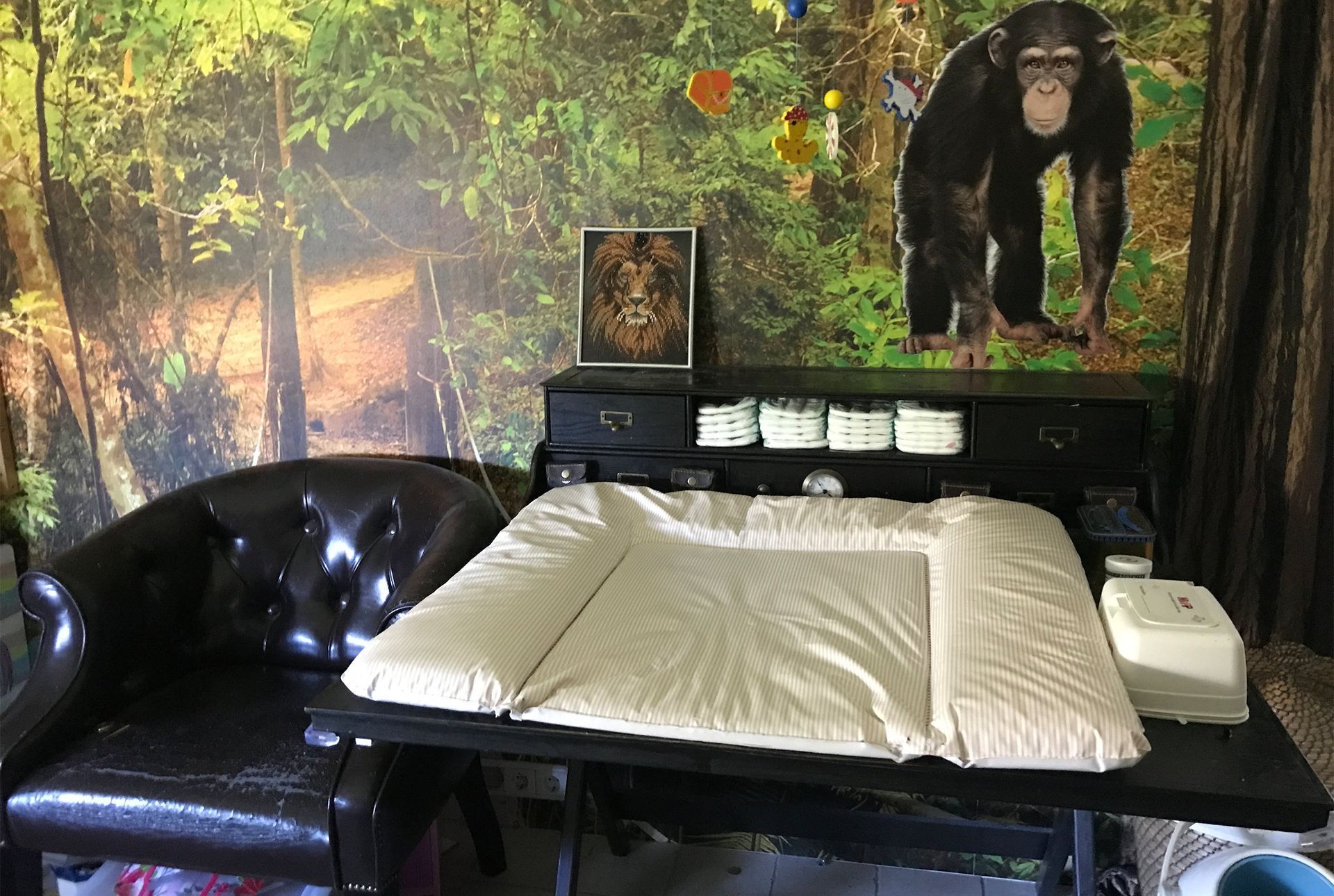 Ein Wickeltisch mit Wickelauflage, umgeben von Dschungel-Dekoration, darunter eine Schimpansen-Figur und ein Ledersessel.