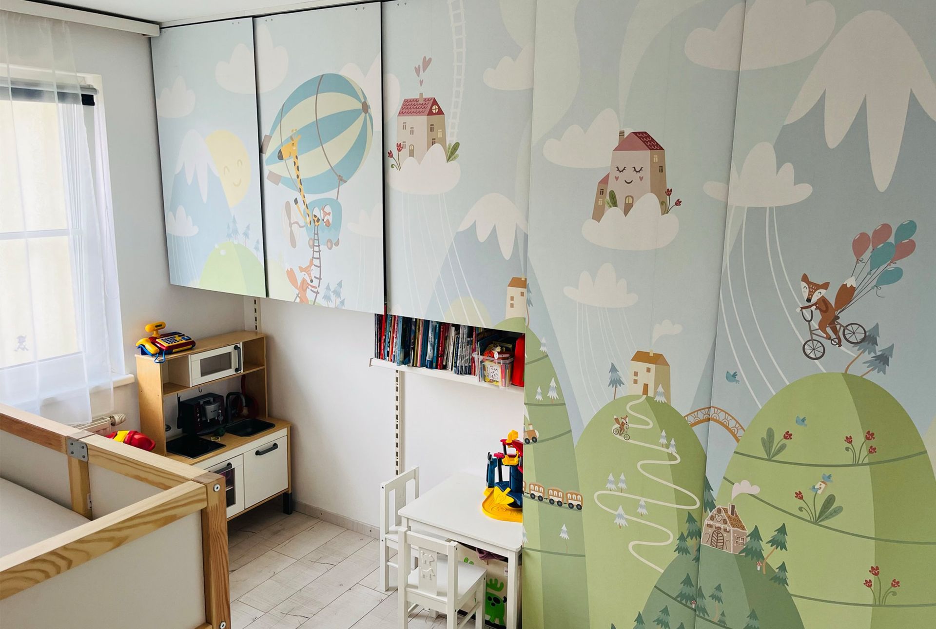 Ein Kinderzimmer mit einer Tapete mit Cartoonmotiven wie Häusern, Bergen und Heißluftballons. Es verfügt über ein Kinderbett und eine Spielküche.