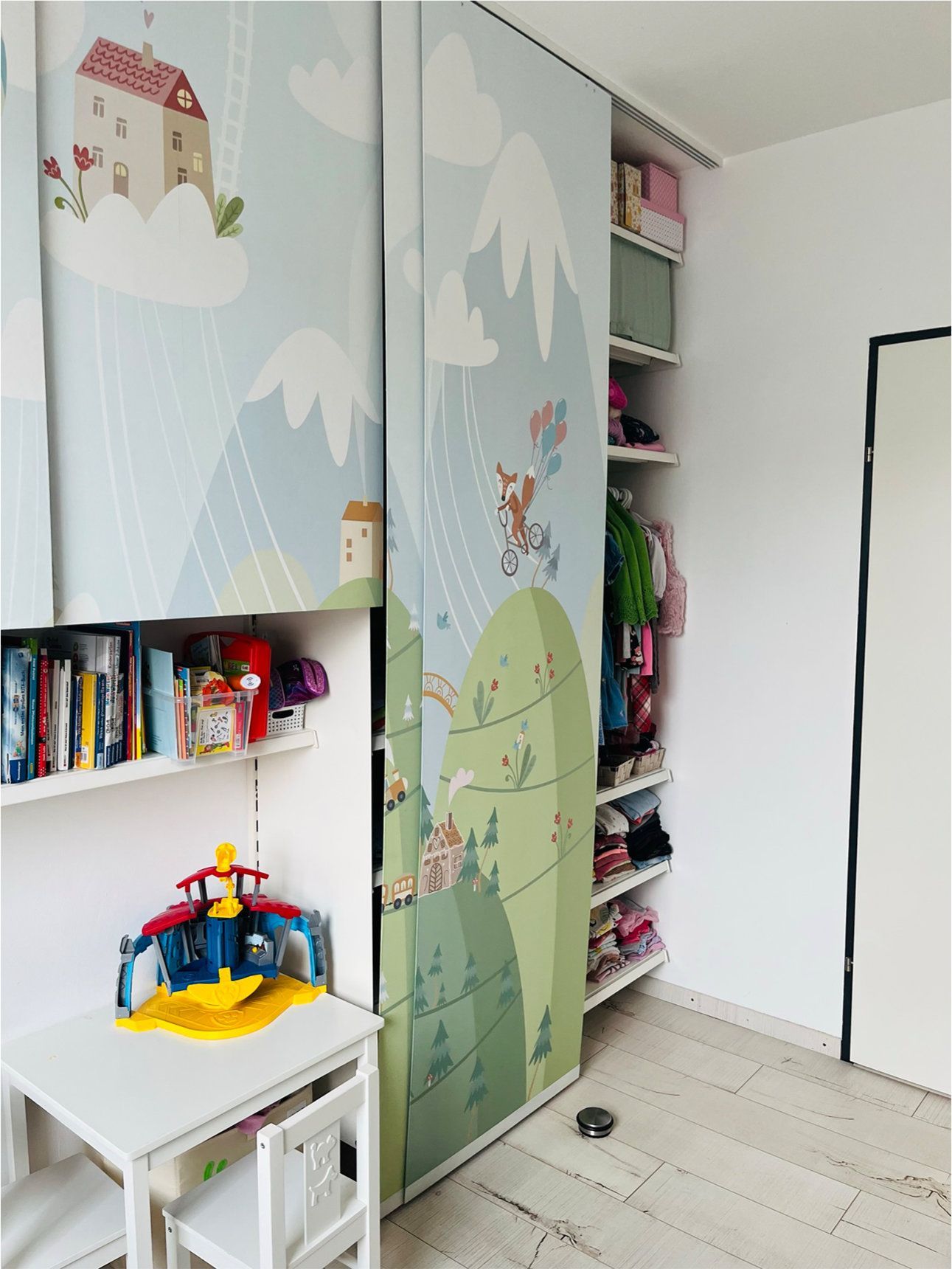 Kinderzimmer mit Berg- und Hausmotiv an den Schranktüren. Bücherregal, Tisch und Spielzeug sind zu sehen.