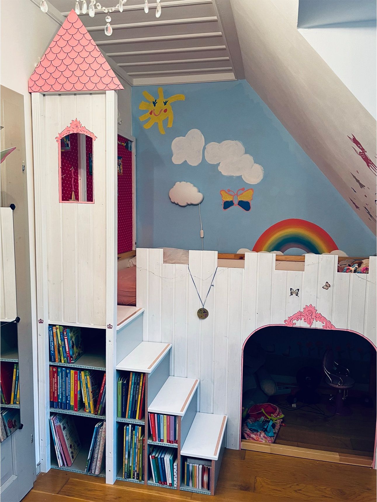 Kinderzimmer mit einem handgefertigten, weiß-rosafarbenen Burgspielhaus mit Bücherregalen und Treppe vor einer blauen Wand mit Regenbogen.