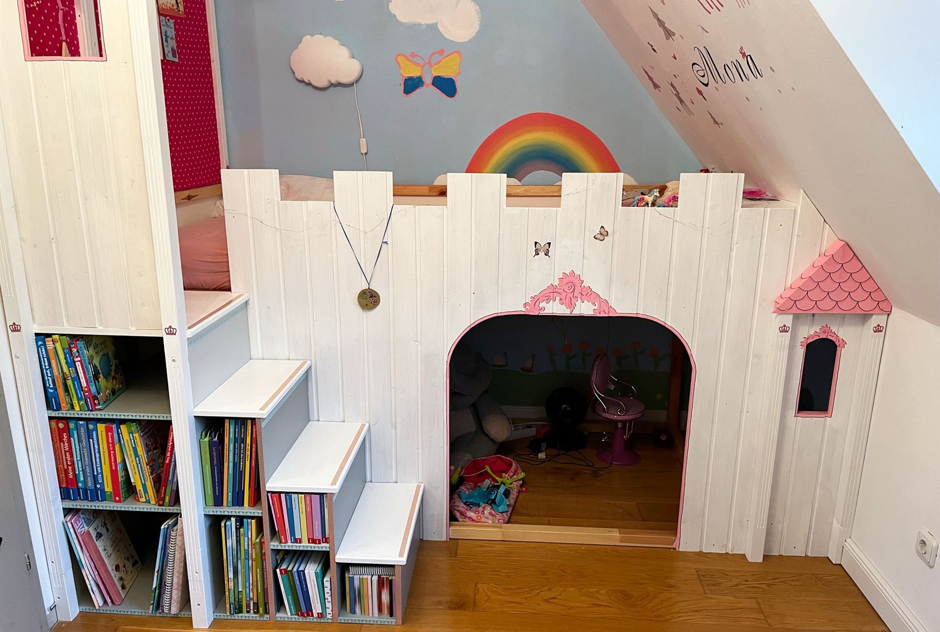 Hochbett im Burgstil im Kinderzimmer mit Treppe und Stauraum, Regenbogen-Wandbild und rosa Akzenten.