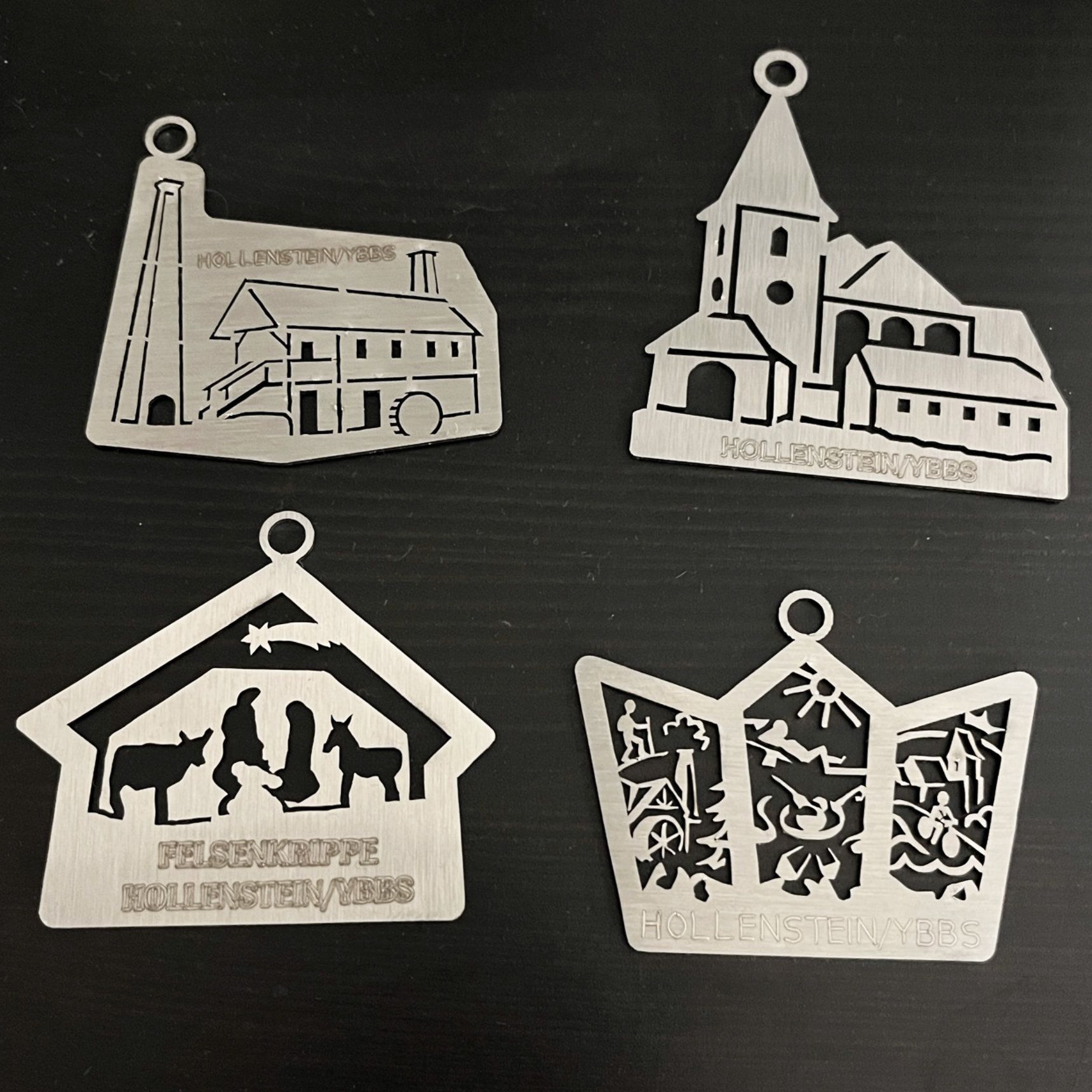 Vier metallische, lasergeschnittene Weihnachtsornamente: Gebäude, Kirche, Krippe und eine verzierte Krone.