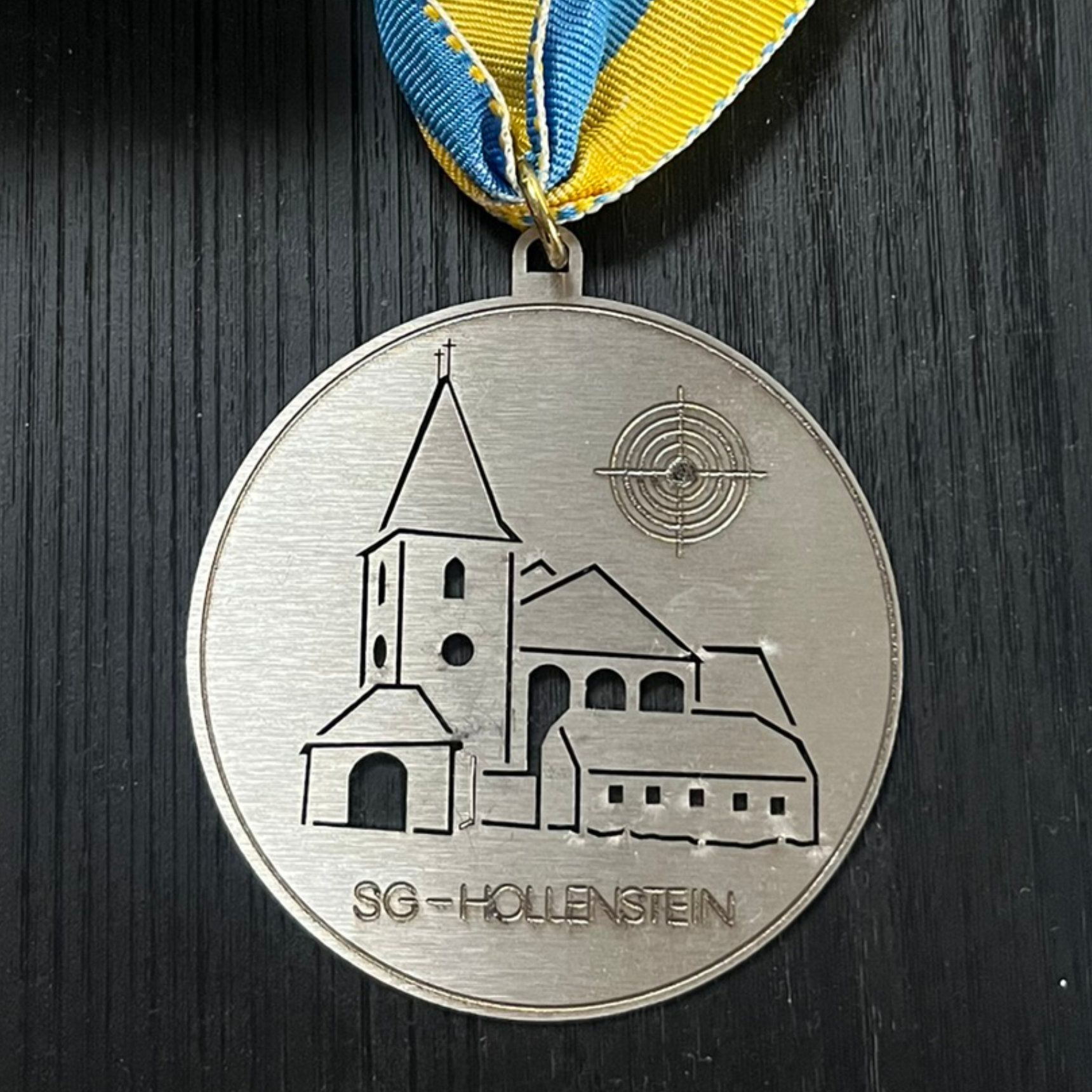 Silbermedaille mit Kirchensilhouette, Zielscheibe und dem Schriftzug „SG-Hollenstein“ auf einem blau-gelben Band.