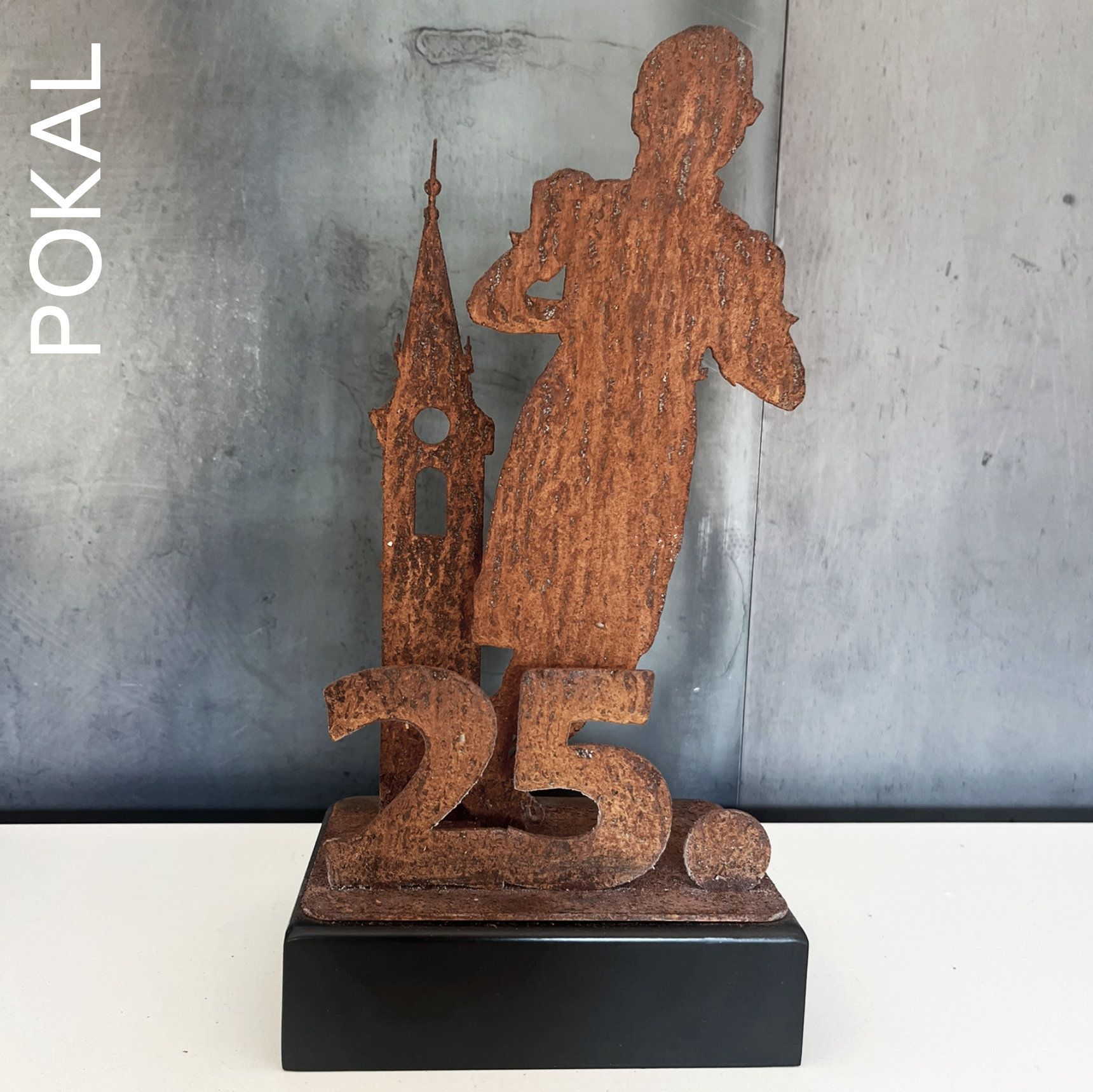 Rostige Metalltrophäe mit einer Figur, einem Turm und der Zahl „25“ auf schwarzem Sockel.
