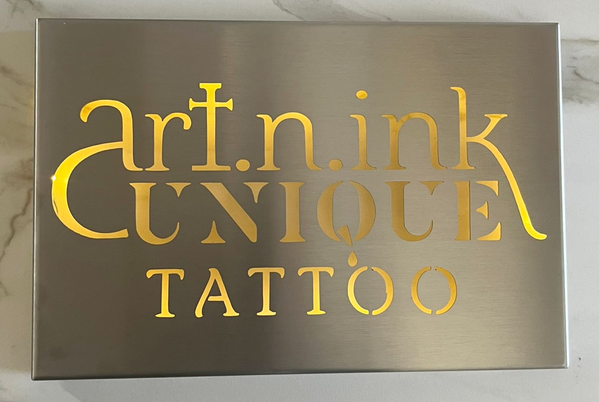 Schild aus Edelstahl mit goldener Aufschrift „art.n.ink UNIQUE TATTOO“.