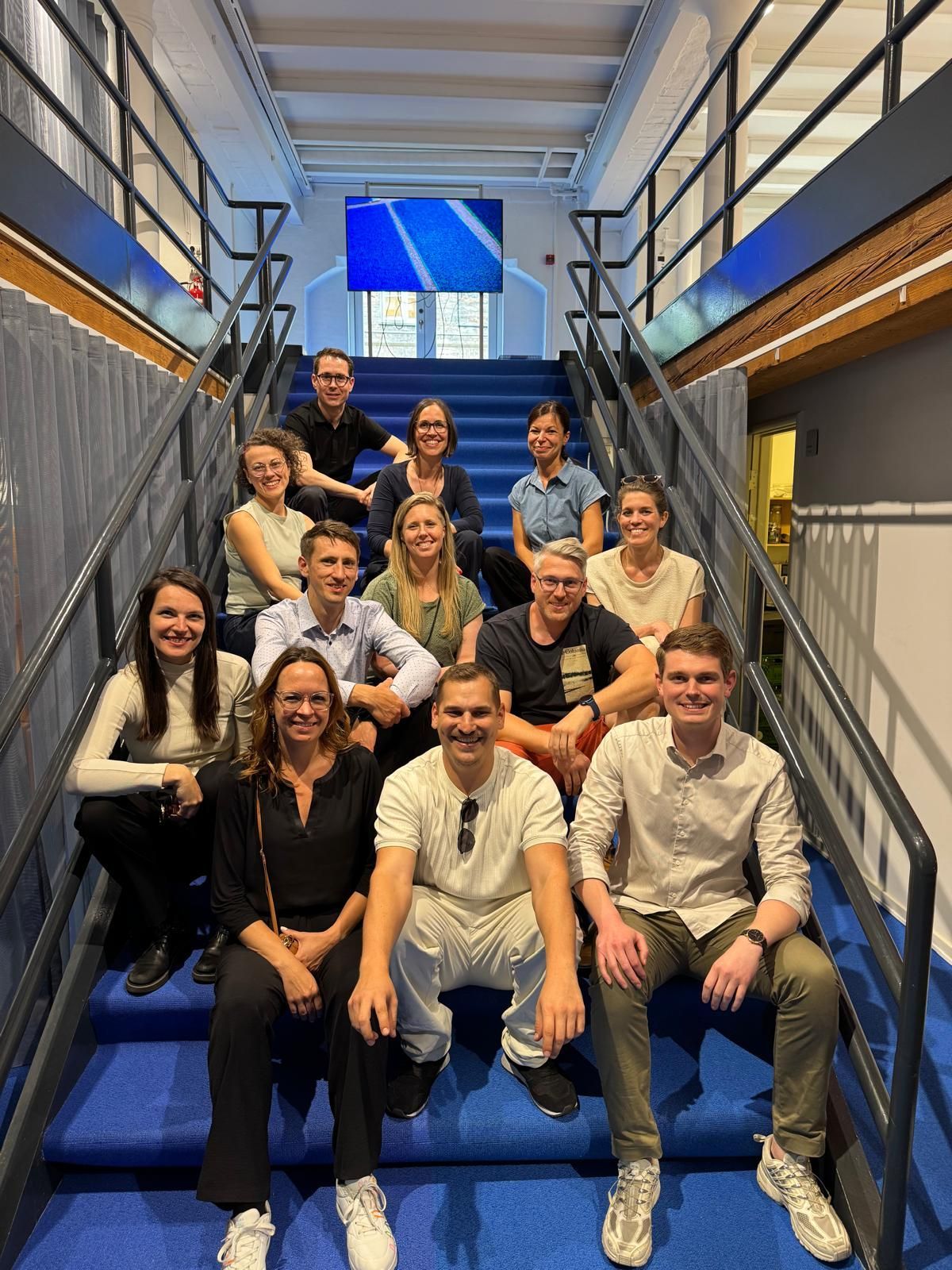 Eine Gruppe von Menschen posiert auf einer blauen Treppe. Innenansicht des Gebäudes mit blauen und weißen Akzenten.