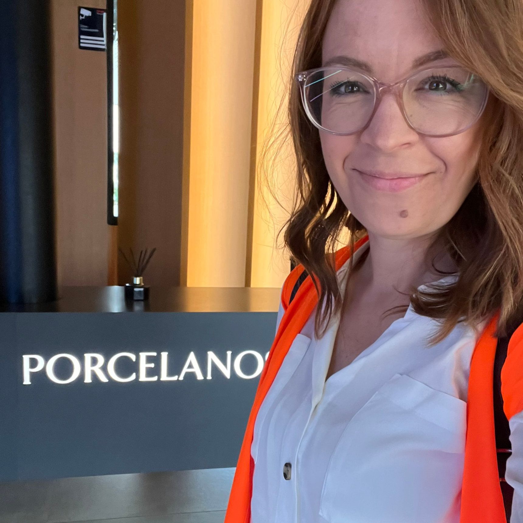 Eine Frau mit Brille lächelt neben einem „PORCELANO“-Schild. Sie trägt ein weißes Hemd und eine orangefarbene Jacke.