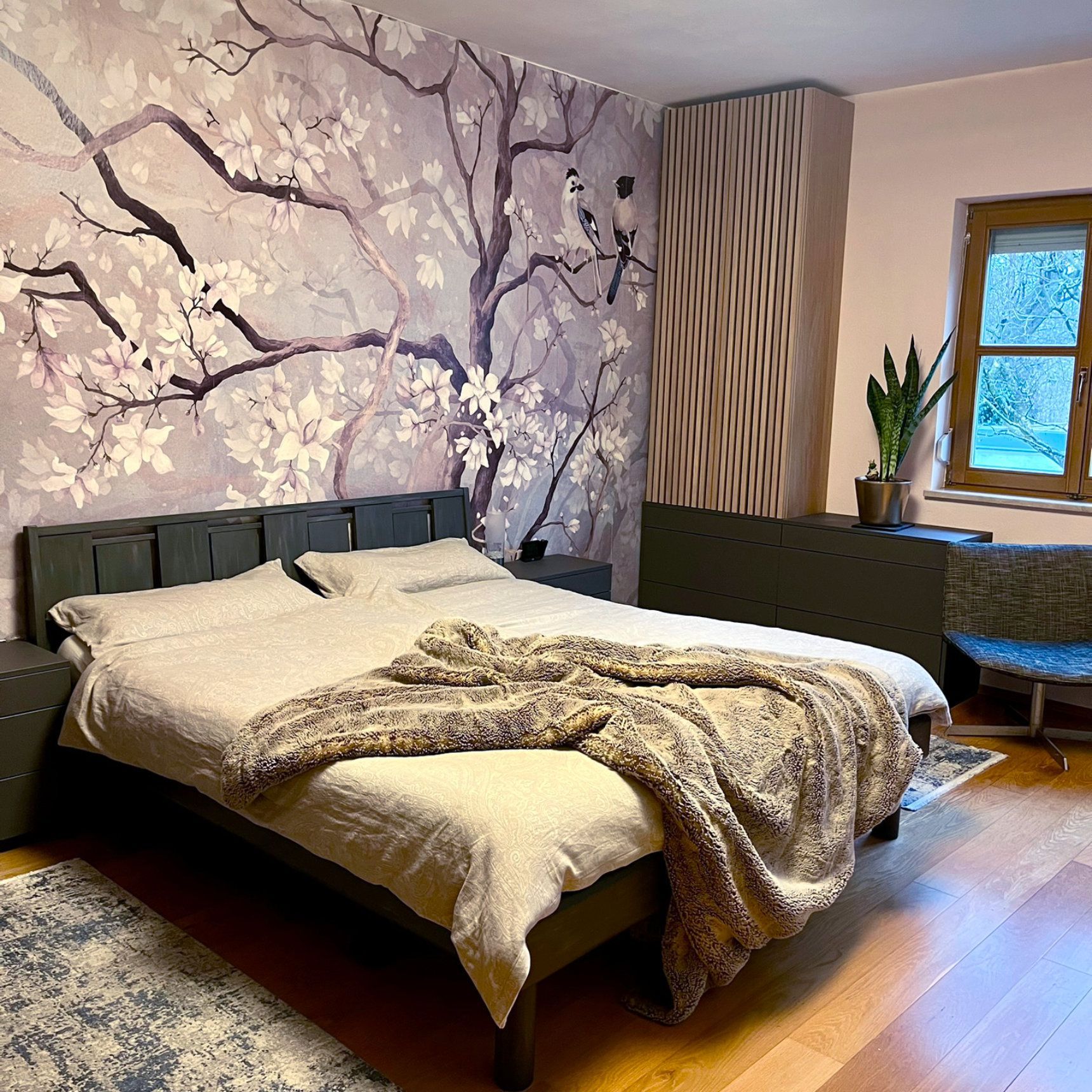 Schlafzimmer mit floralem Wandbild, grauem Bett, Holzboden und Fenster.