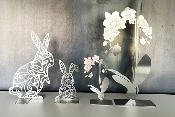 Lasergeschnittene Osterdekorationen aus Metall: Hasen und Orchideenblüten auf weißem Grund vor grauem Hintergrund.