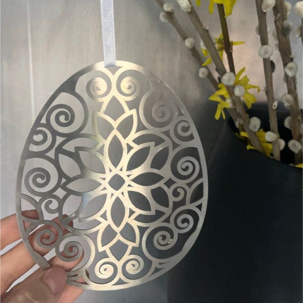Ein dekoratives Osterei aus Metall mit floralen Ausschnitten wird von einer Hand gehalten. Im Hintergrund ist eine Vase mit gelben Blumen zu sehen.