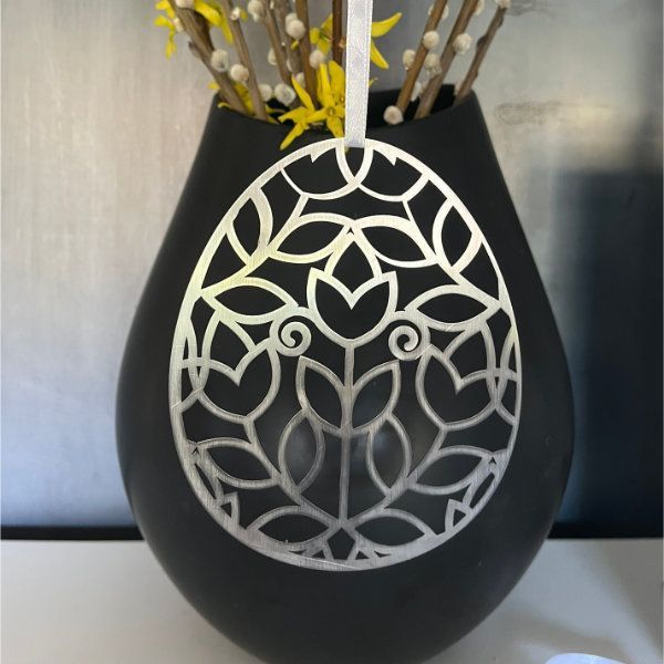 Ein silbernes Osterei mit Blumenmuster hängt vor einer schwarzen Vase mit gelben Blumen.