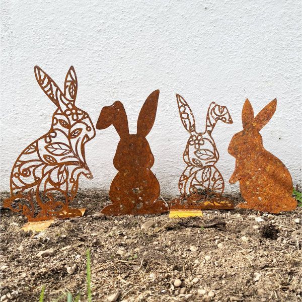 Vier rostige Metallhasen-Silhouetten stehen in einem Gartenbeet.