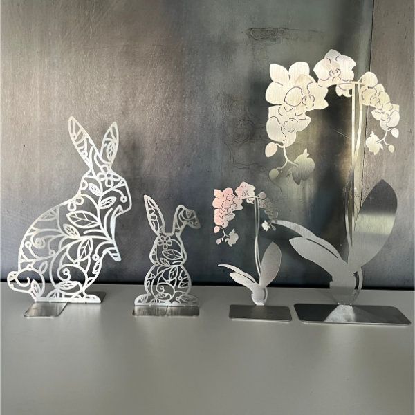 Osterdekorationen aus lasergeschnittenem Metall: zwei Hasen und eine Orchidee mit rosa Blüten auf einem Tisch vor silbernem Hintergrund.