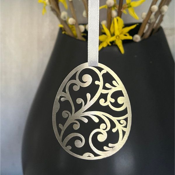 Ein goldener, filigraner Osterei-Anhänger hängt vor einer schwarzen Vase mit gelben Blumen.