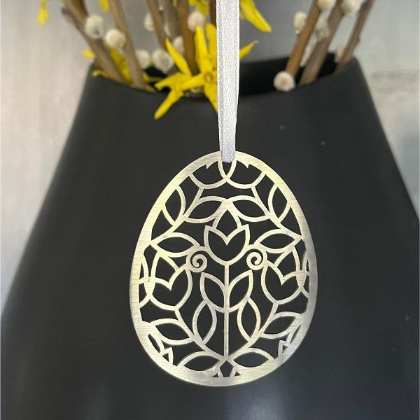 Goldfarbenes, lasergeschnittenes Eierornament mit floralem Muster, das an einem weißen Band hängt, vor einer dunklen Vase mit gelben Blumen.